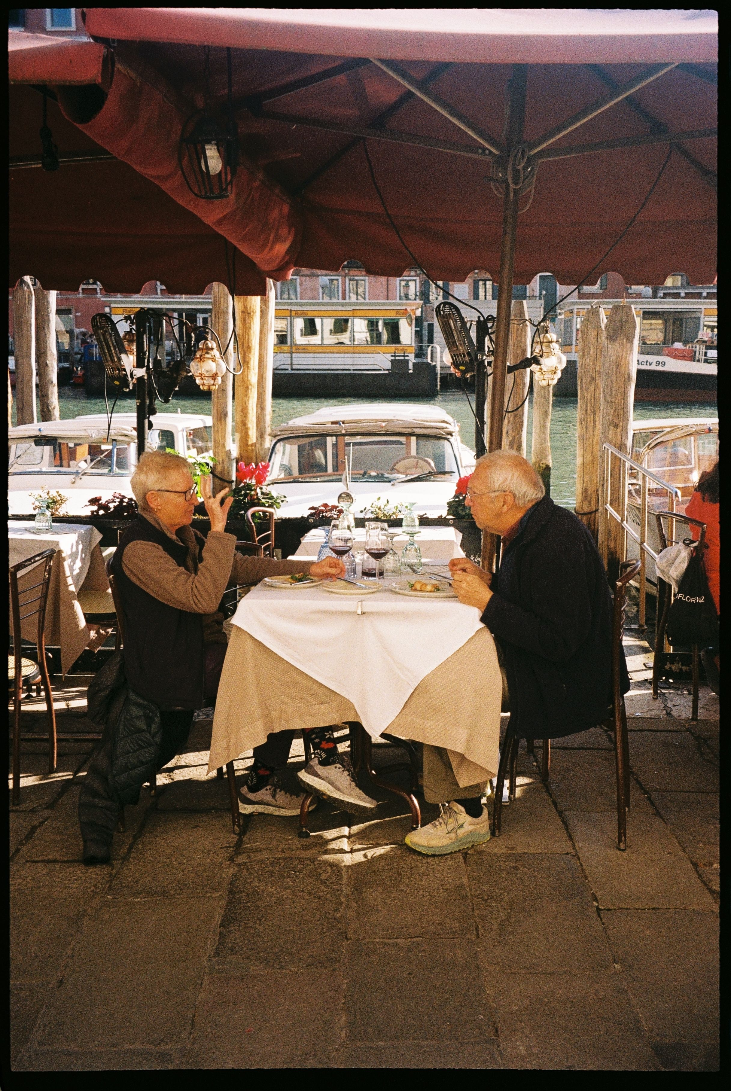 Two elderly men dining at a cafe by the Venice canal, enjoying a sunny day.
