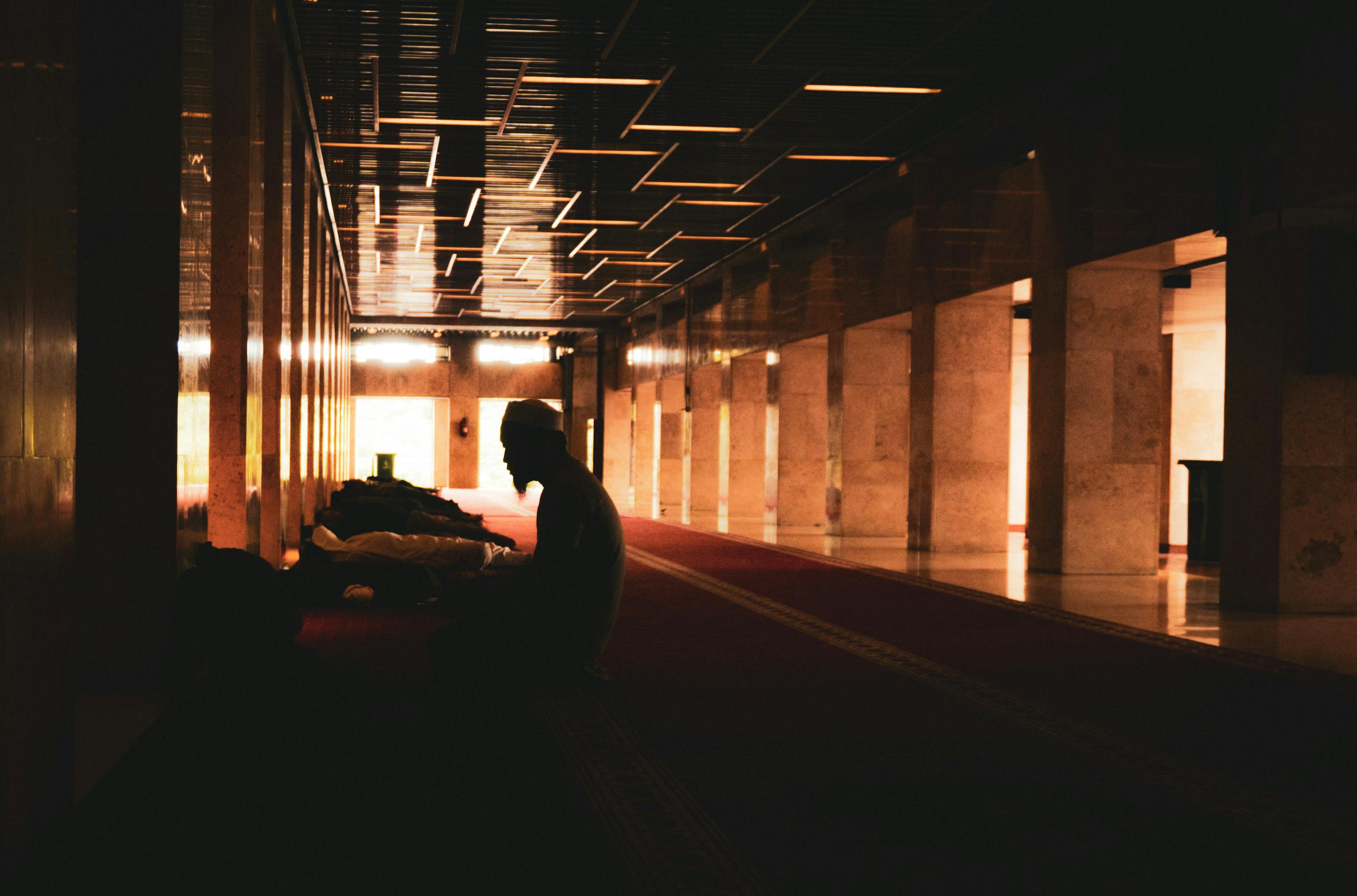 Gratis En silhuett av en person som ber inne i en svakt opplyst moské med reflekterende tak og søyler. Arkivbilde