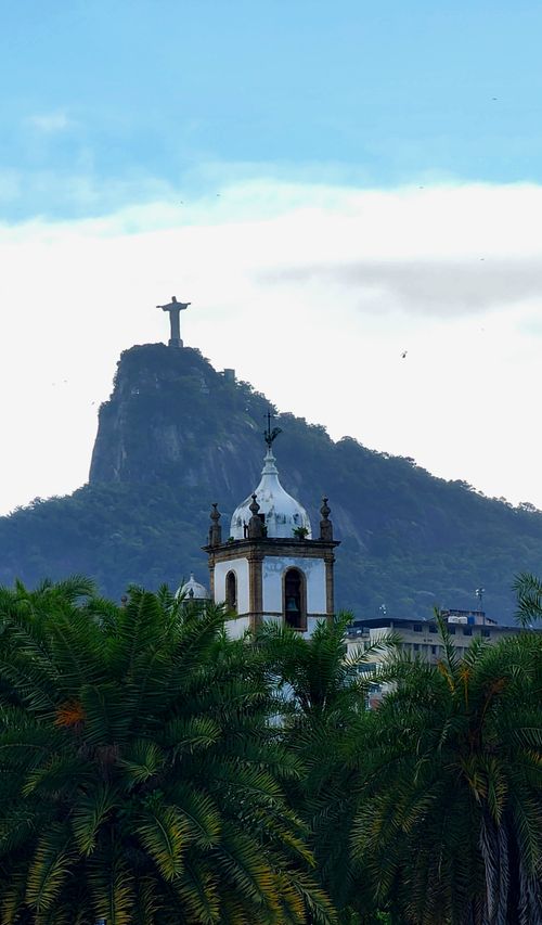 Free Dramatic view of Christ the Redeemer statue from afar, with palm trees and a historic building in the foreground. Stock Photo
