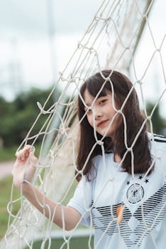 Young woman in soccer jersey smiling through a goal net outdoors.