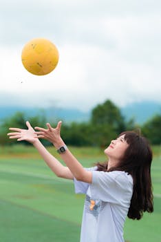 Young woman wearing a white shirt plays soccer outdoors, catching a yellow ball with a joyful expression.