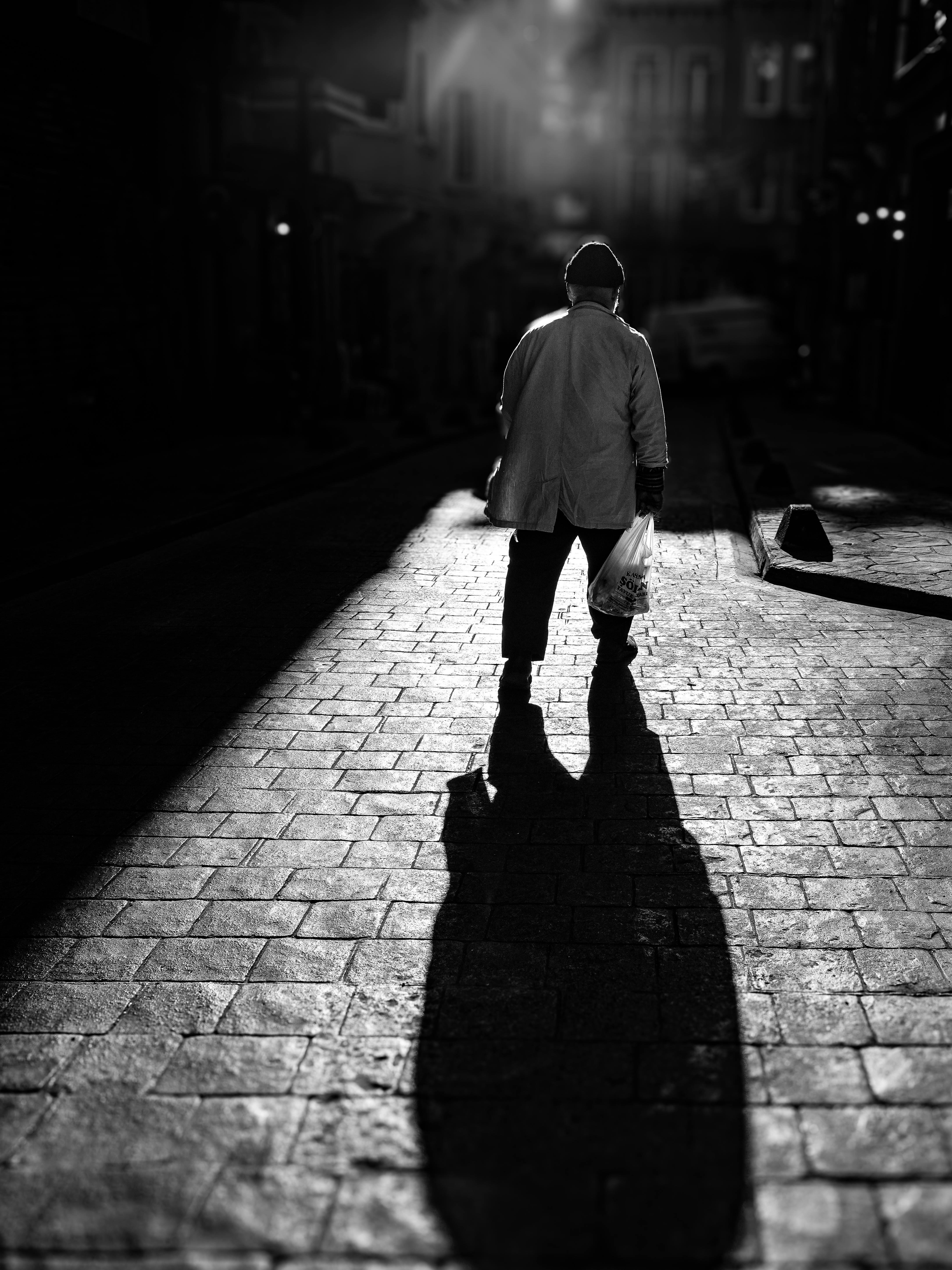 Gratuit O siluetă izbitoare în alb-negru a unei persoane care merge pe o stradă pietruită și goală, aruncând o umbră lungă. Fotografie de stoc