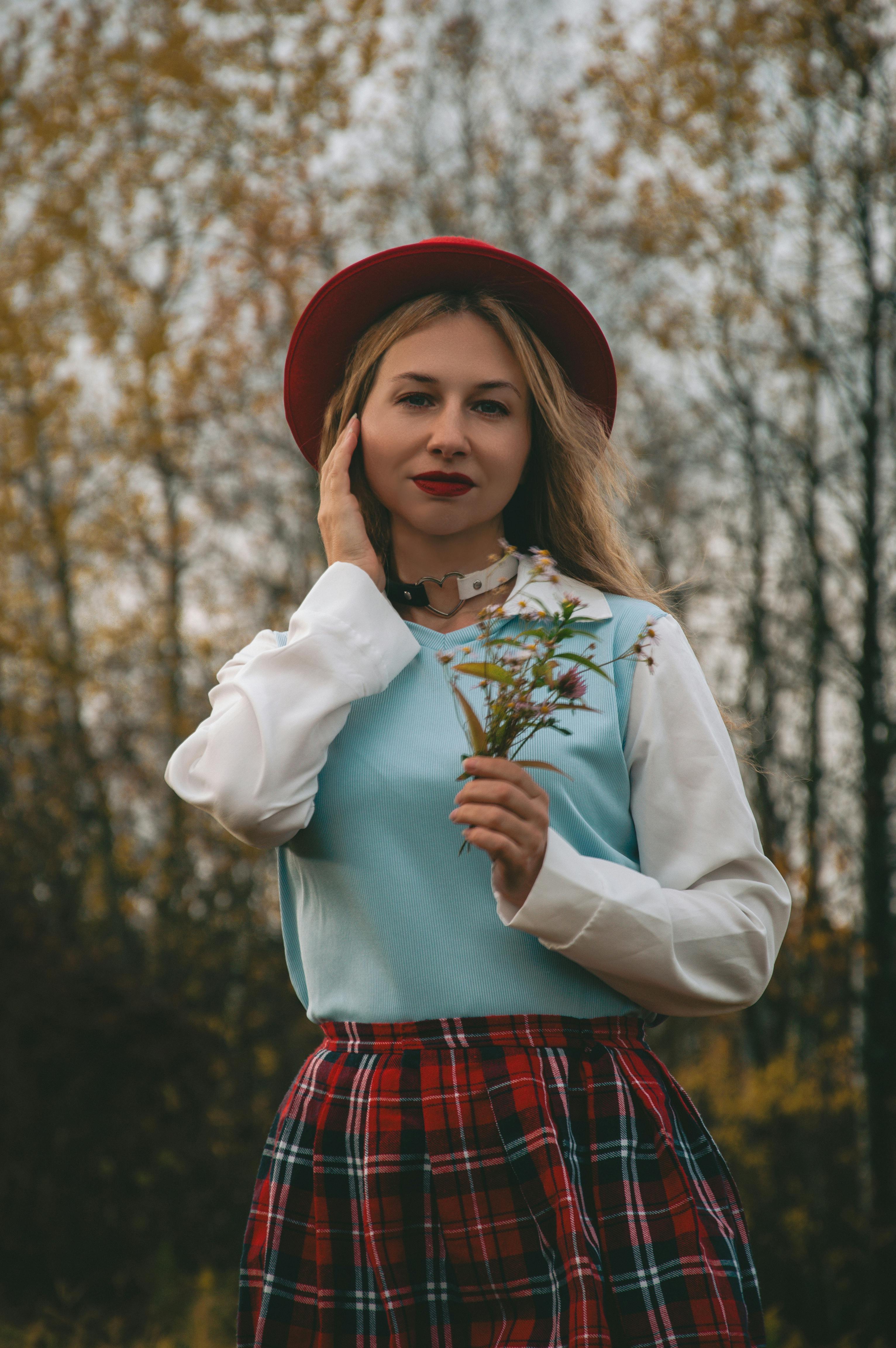 Gratis Una donna alla moda con un cappello rosso tiene in mano dei fiori selvatici all'aperto in un ambiente autunnale. Foto a disposizione
