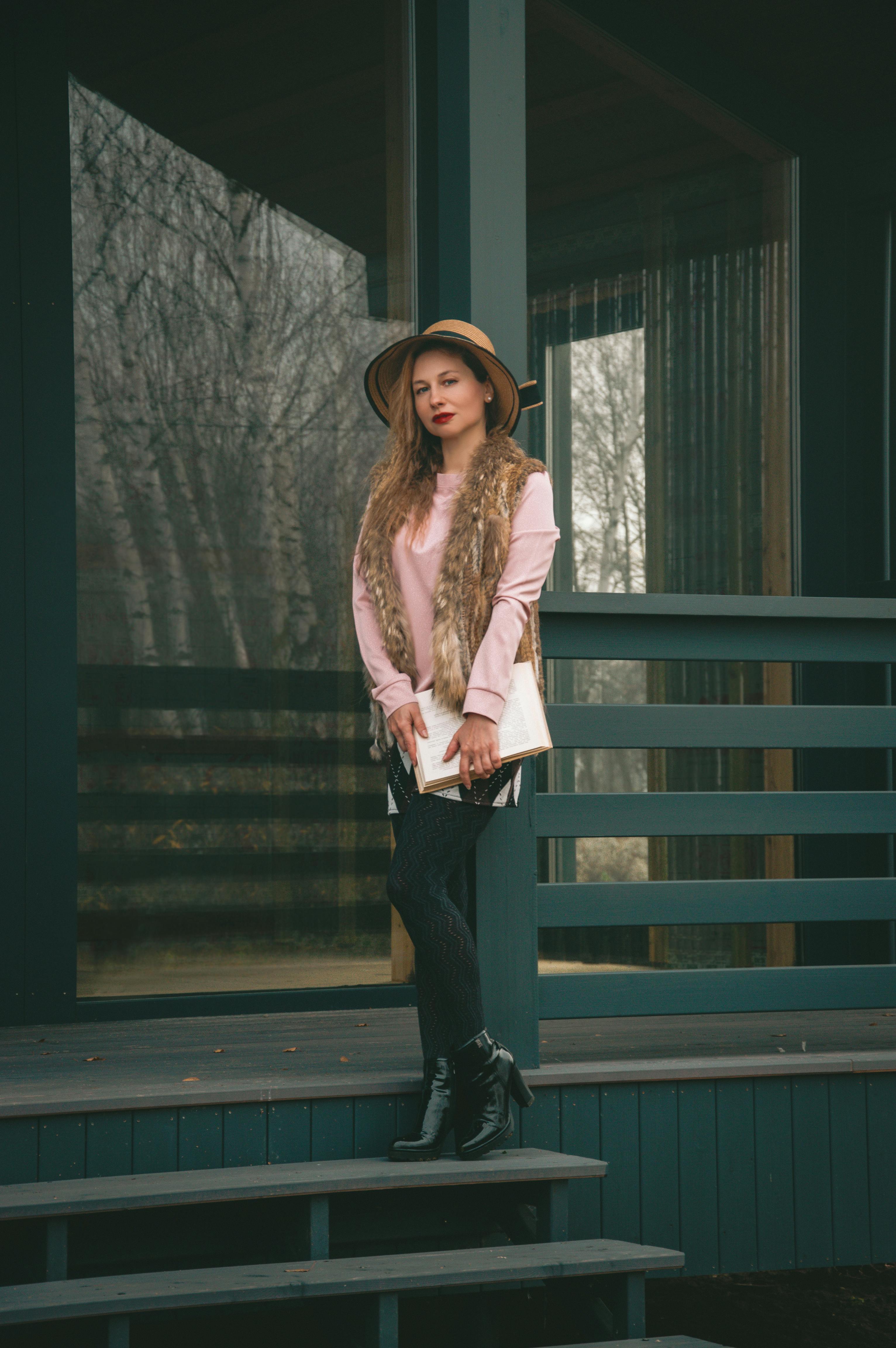 무료 모자와 모피 조끼를 입은 세련된 여성이 나무 오두막 계단에 앉아 포즈를 취하고 있다. 스톡 사진