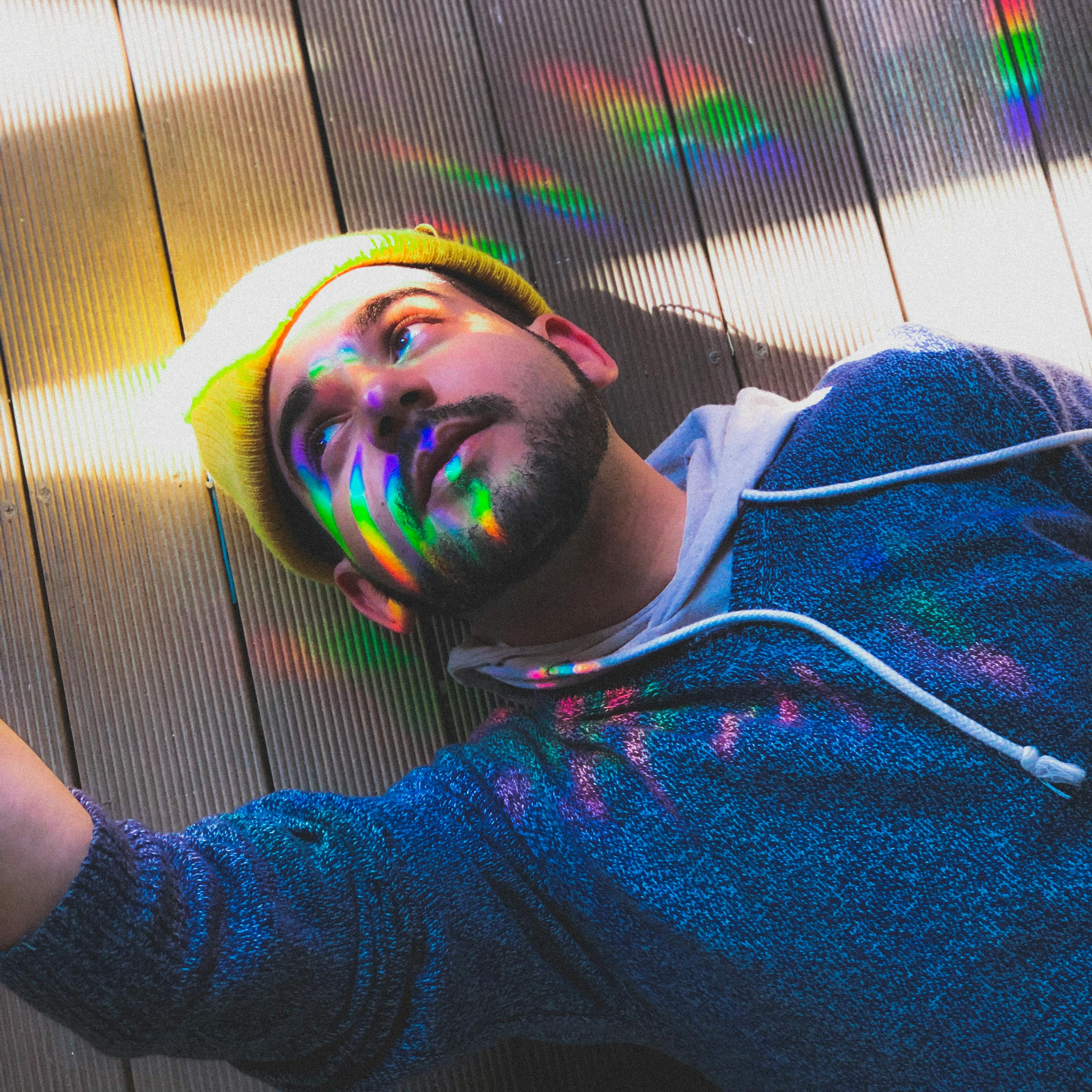 在户外木制甲板上，一位留着胡须、穿着连帽衫的时尚男子被彩虹般的灯光照亮