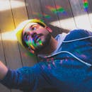 在户外木制甲板上，一位留着胡须、穿着连帽衫的时尚男子被彩虹般的灯光照亮