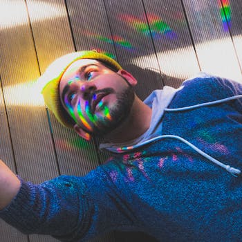 在户外木制甲板上，一位留着胡须、穿着连帽衫的时尚男子被彩虹般的灯光照亮