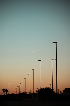 Street lamps in silhouette line a sunset sky with a distant airplane. Perfect for serene and urban-themed designs.