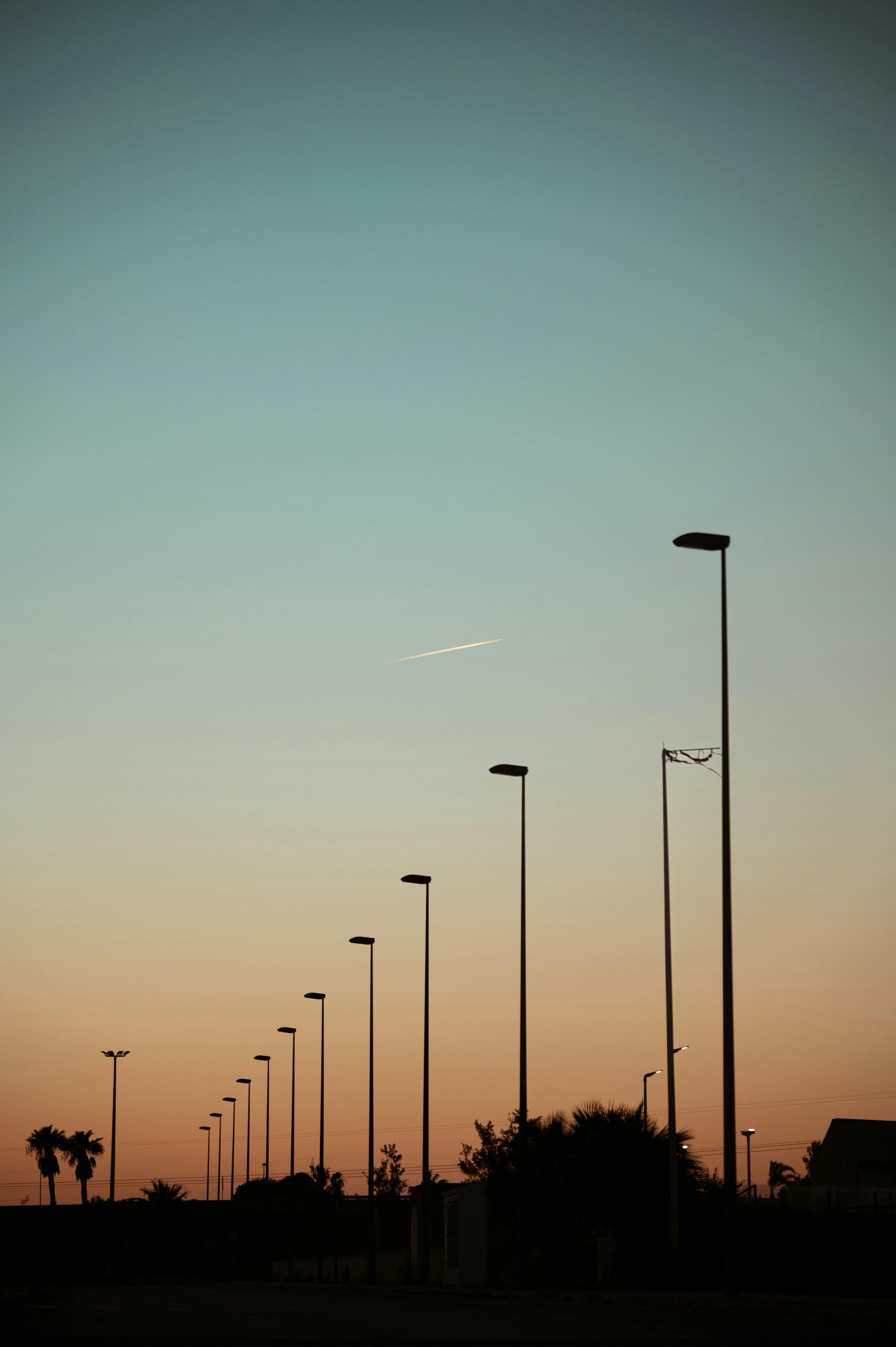 Kostenlos Straßenlaternen zeichnen sich als Silhouetten vor einem Sonnenuntergangshimmel ab, in der Ferne ist ein Flugzeug zu sehen. Perfekt für ruhige und urbane Designs. Stock-Foto