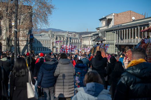 A vibrant street parade in Tbilisi, with people celebrating in winter attire.