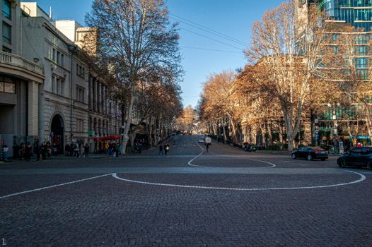 A bustling city street scene in Tbilisi, Georgia, with fall foliage and architectural landmarks.