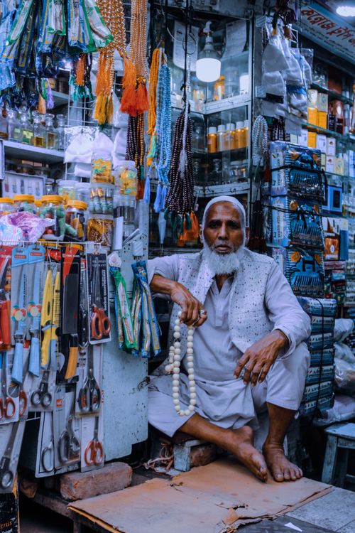 Free A man in traditional attire sits beside a vibrant assortment of market goods, embodying local culture. Stock Photo