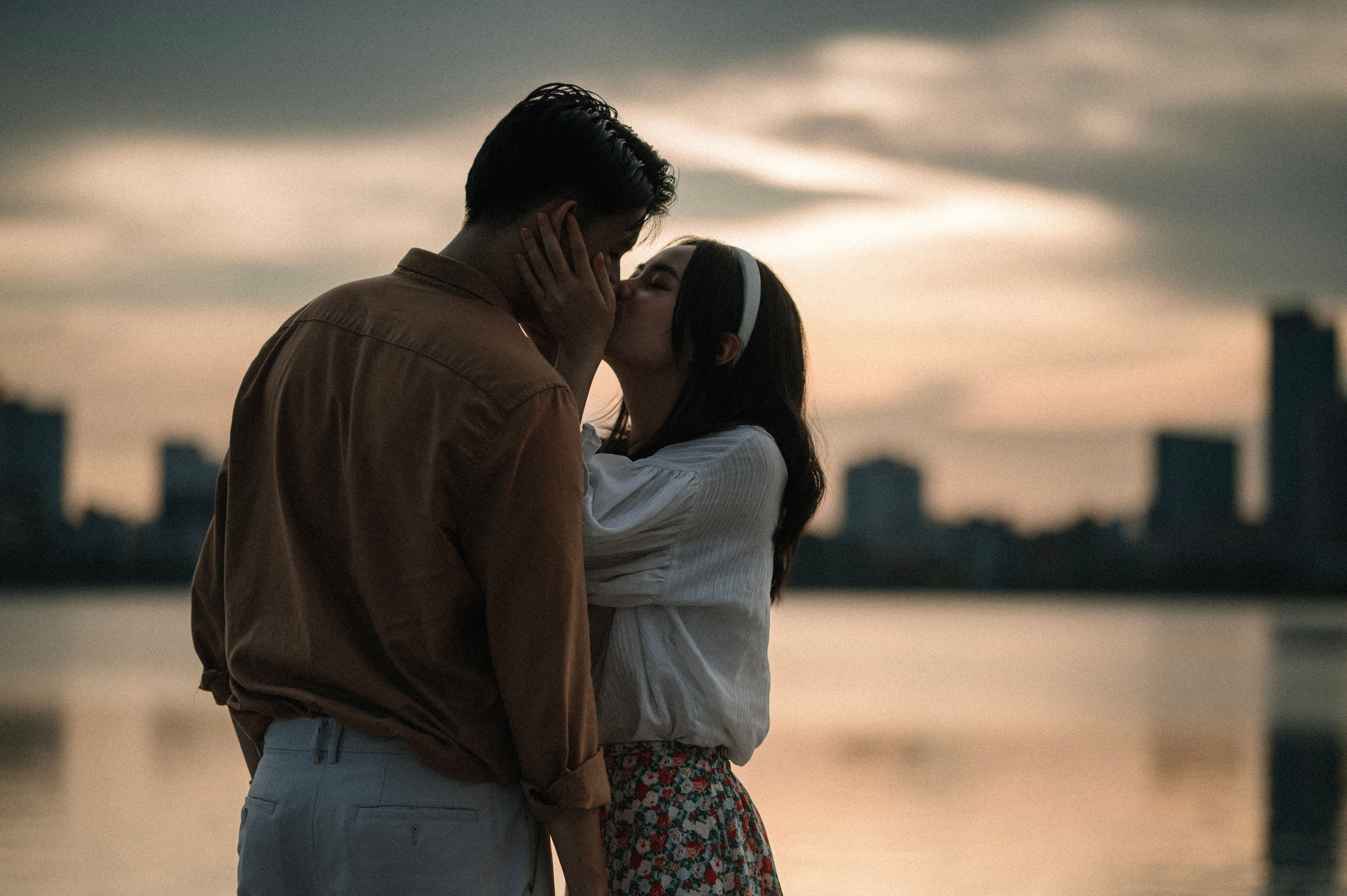 A couple shares a romantic kiss by the lake at sunset with city skyline in the background.