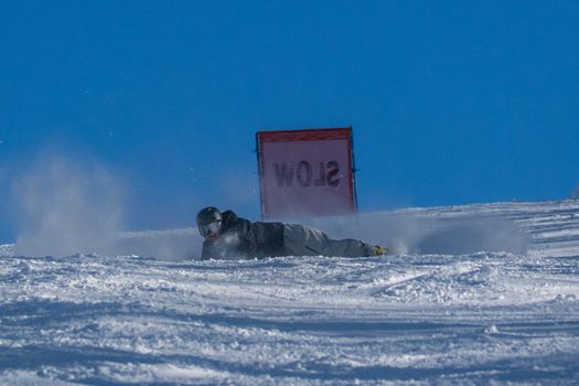 Snowboarder sliding on snow past a 'Slow' sign under a clear blue sky.