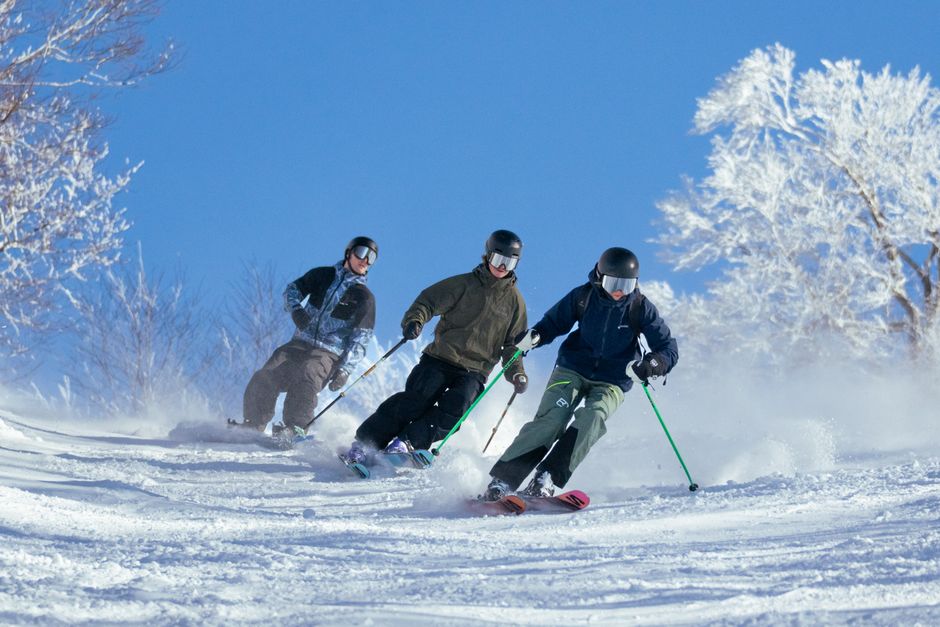 Three skiers carve down a snowy mountain slope on a bright winter day.