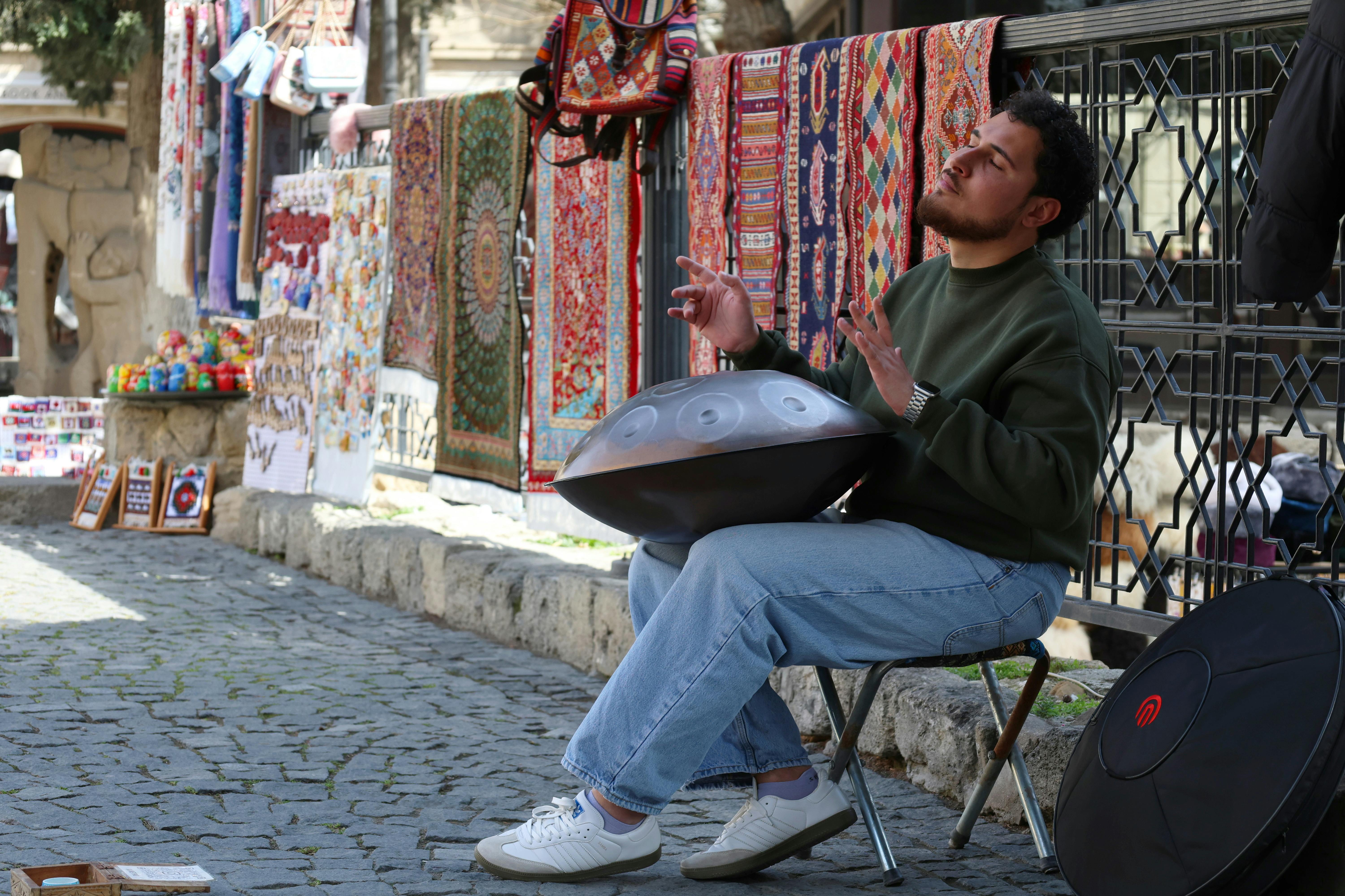 Free A man plays a handpan at a bazaar with vibrant tapestries displayed, capturing urban street culture. Stock Photo