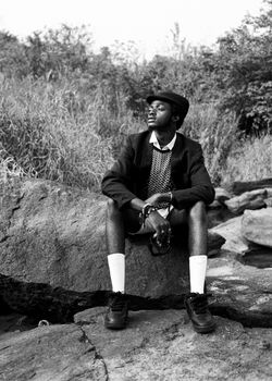 Black and white photo of a stylish man sitting on rocks outdoors, in a contemplative pose.