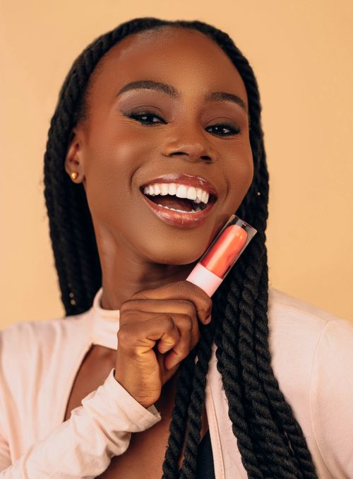 Free Joyful woman with braided hair holding lipstick in a cheerful expression against a beige backdrop. Stock Photo