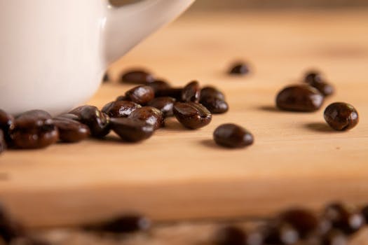 A close-up image of dark roasted coffee beans scattered on a wooden surface with a coffee mug in the background.