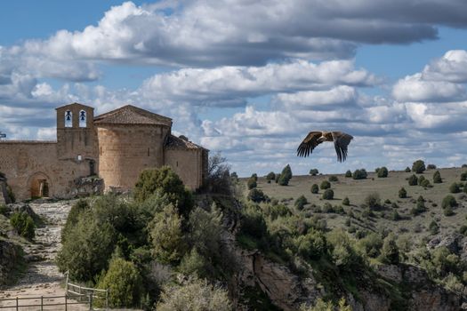 A vulture soars over a historic church in Sepúlveda, Spain, under a dramatic sky.
