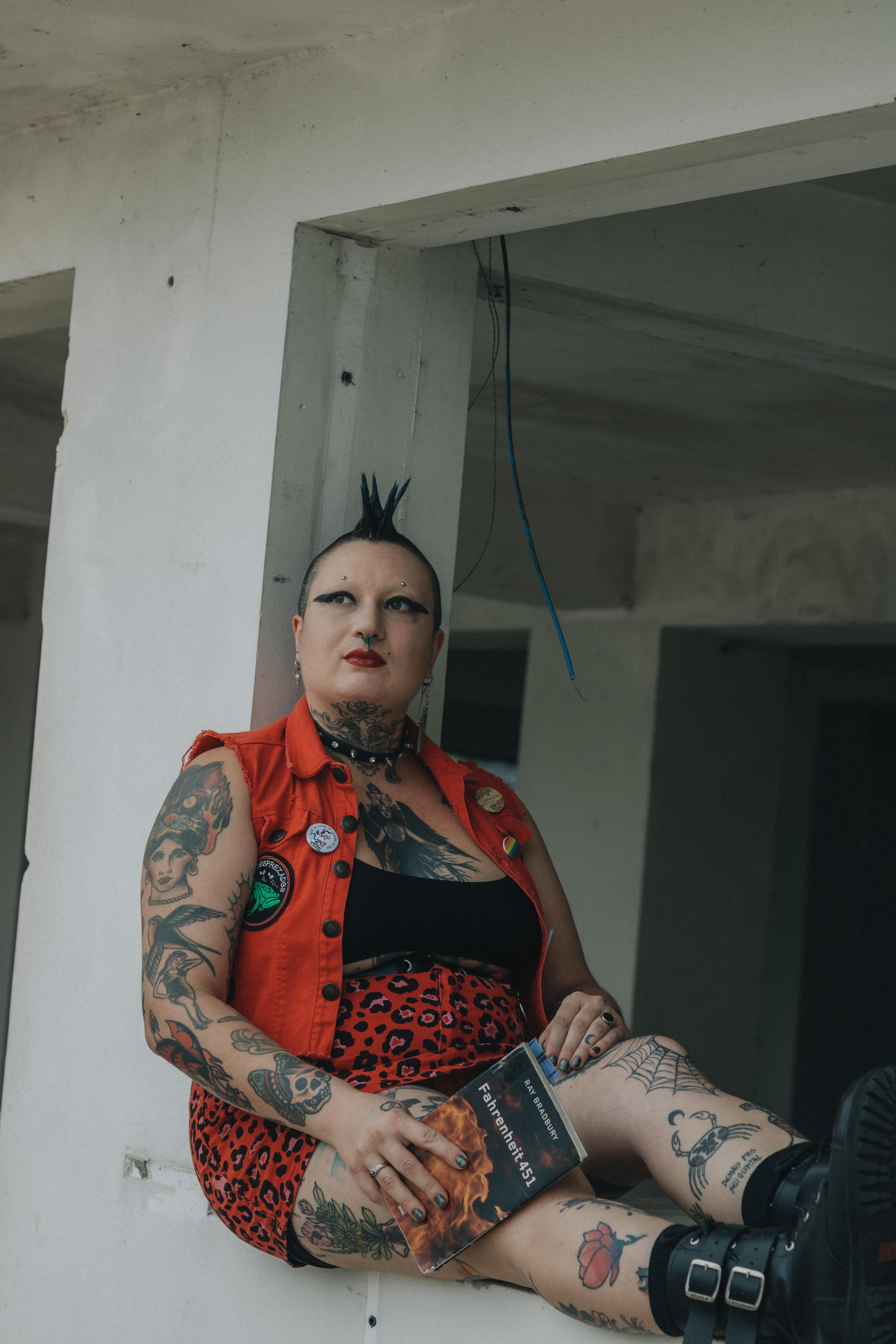 Безкоштовне Панк-жінка з татуюваннями та ірокезом сидить у приміщенні та тримає книгу. стокове фото