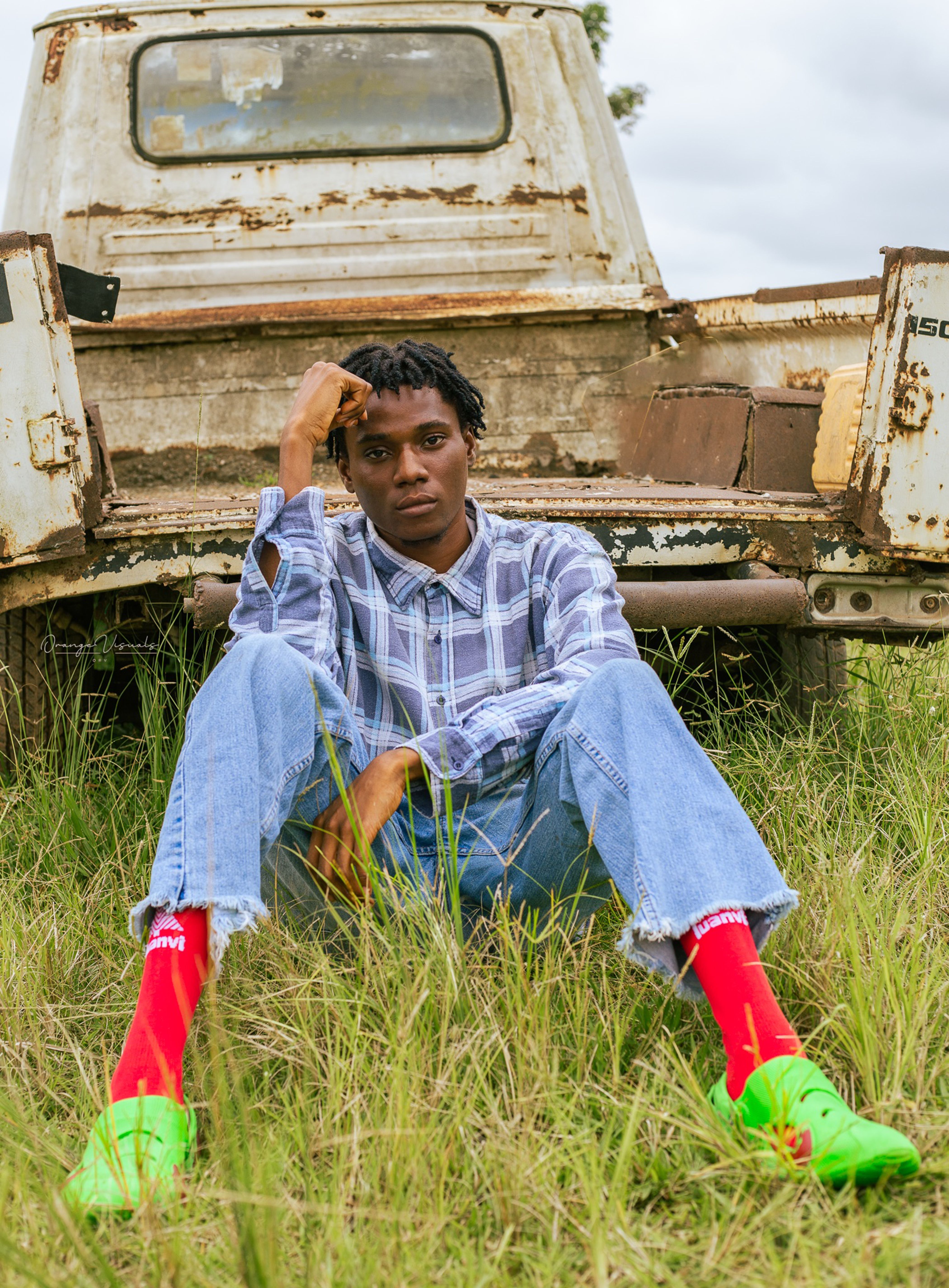 Young man in casual outfit sitting against a rustic pickup truck in a grassy field.