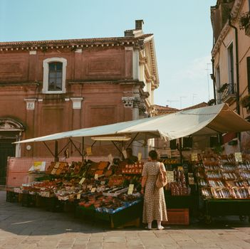 A serene scene of a market stall in Venice, Italy, with a woman browsing the fresh produce.