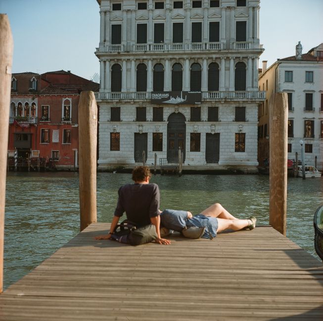 Couple lounging on a Venetian dock, with grand architecture in the background.