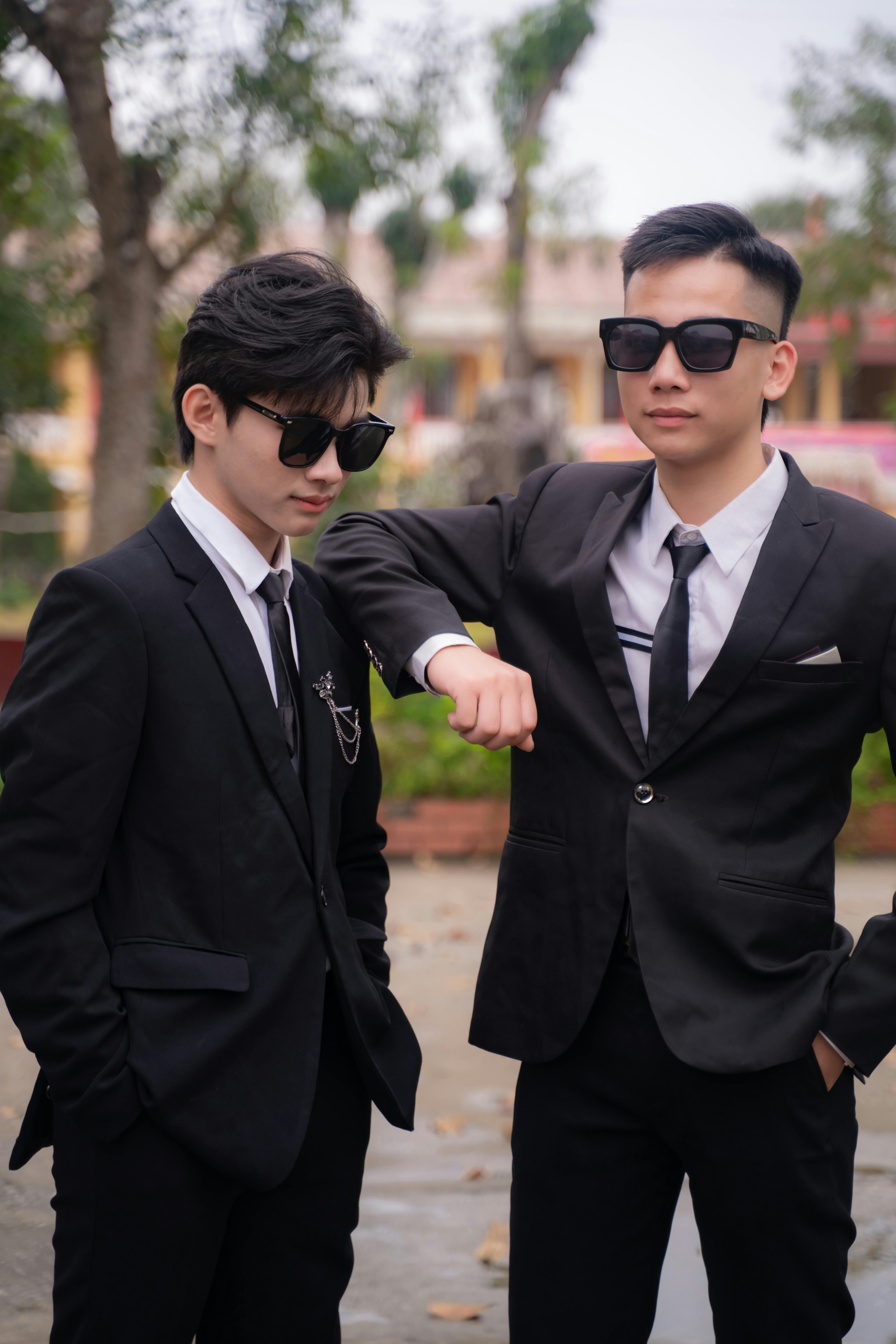 無料 黒いスーツにサングラスをかけた二人の若い男性が、屋外で自信満々にポーズをとっている。 写真素材