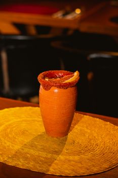 A clay cup with rim garnished with chili and an orange slice on a woven mat.