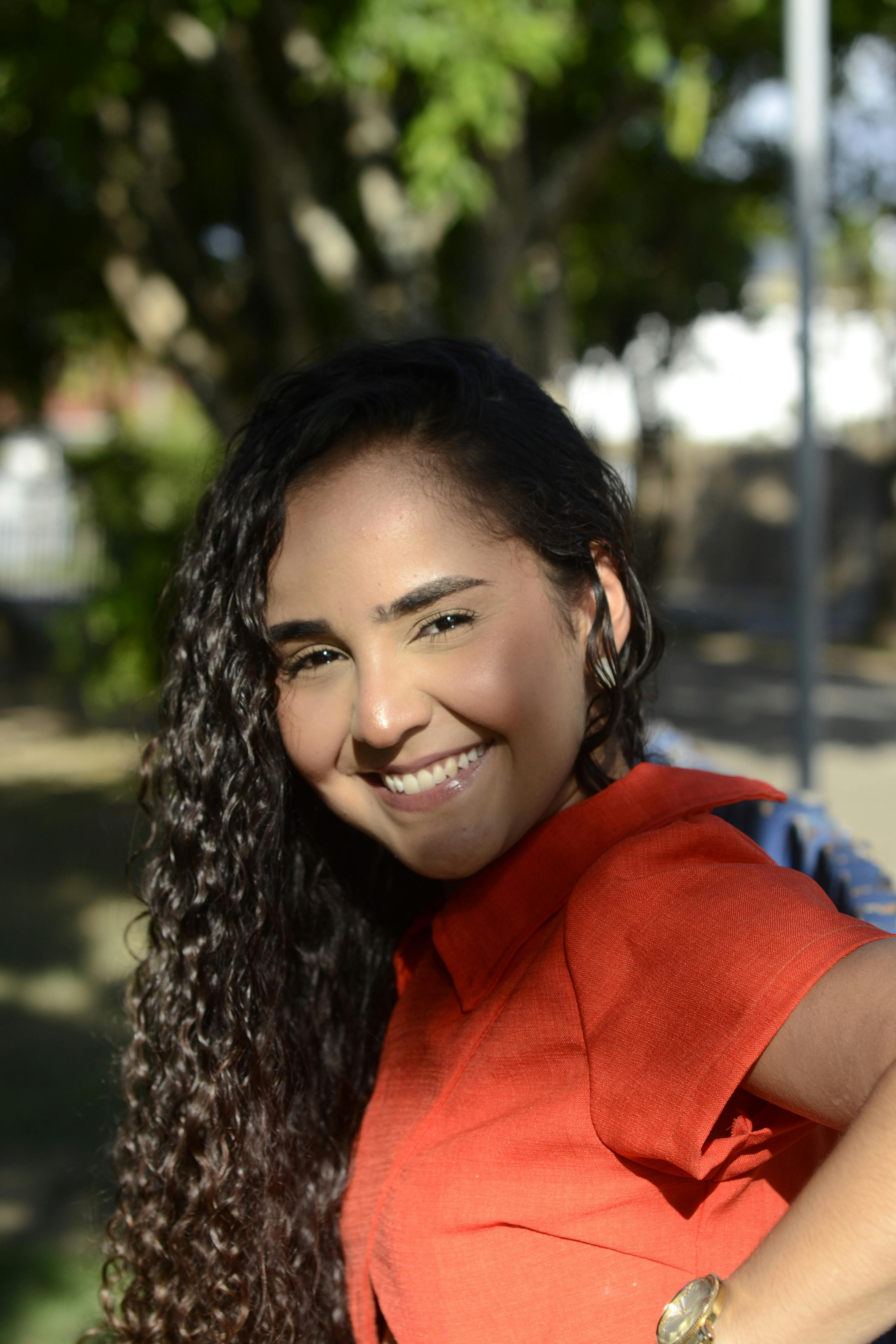 Portrait of a smiling woman with curly hair in a sunlit park setting, wearing a red shirt.