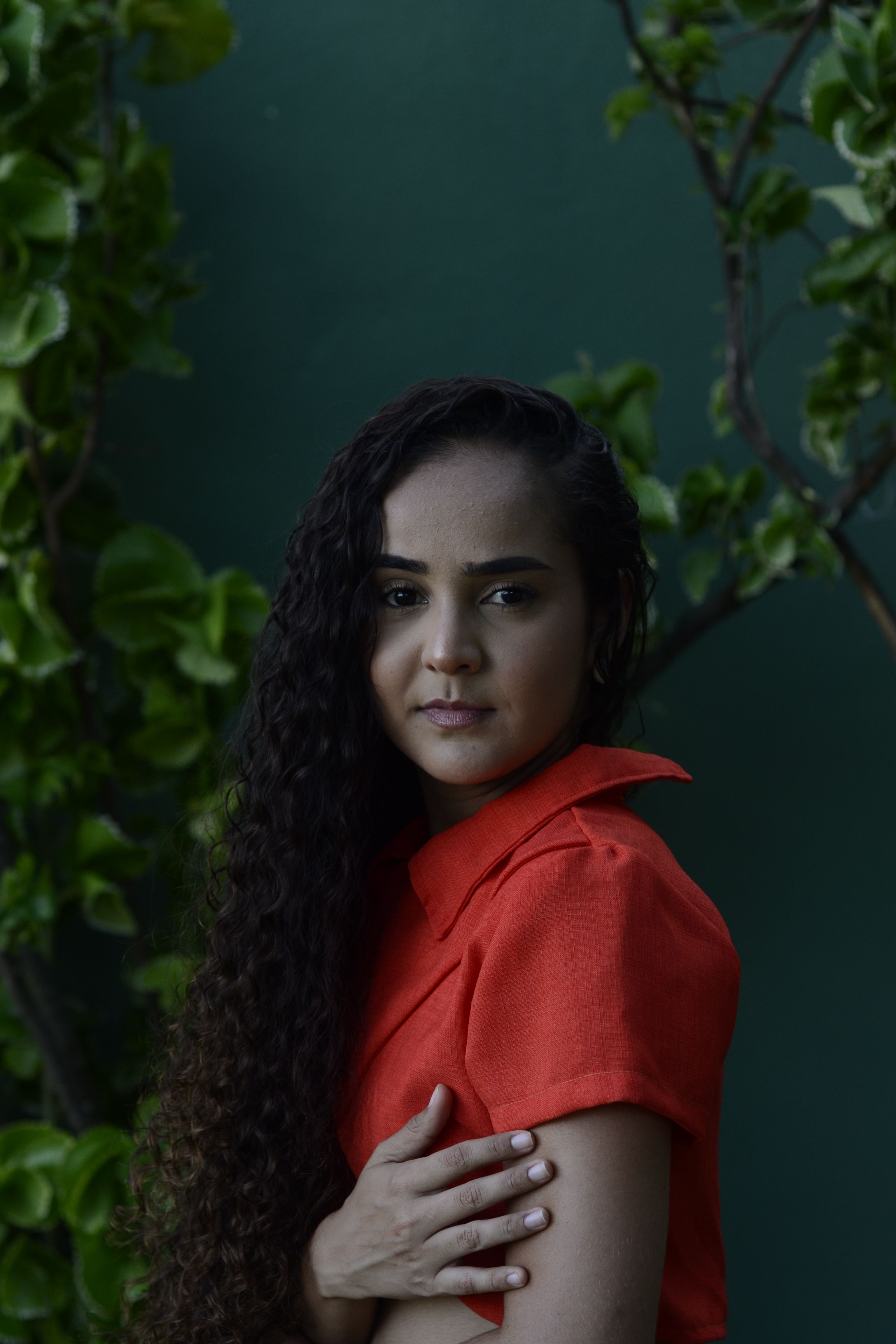 A young woman with curly hair poses against a green leafy background outdoors.