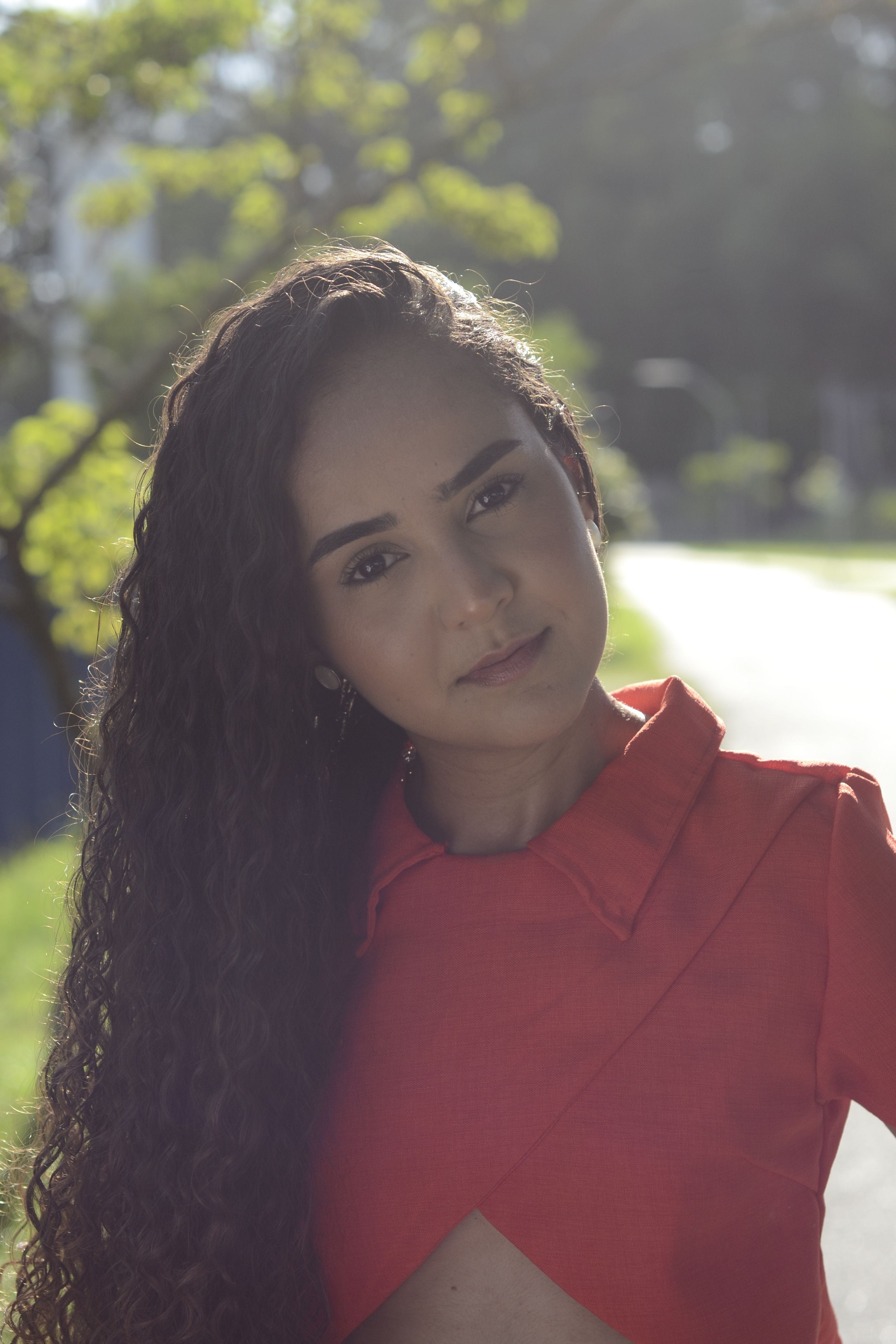 Woman with long curly hair in vibrant red top, serene outdoor portrait.