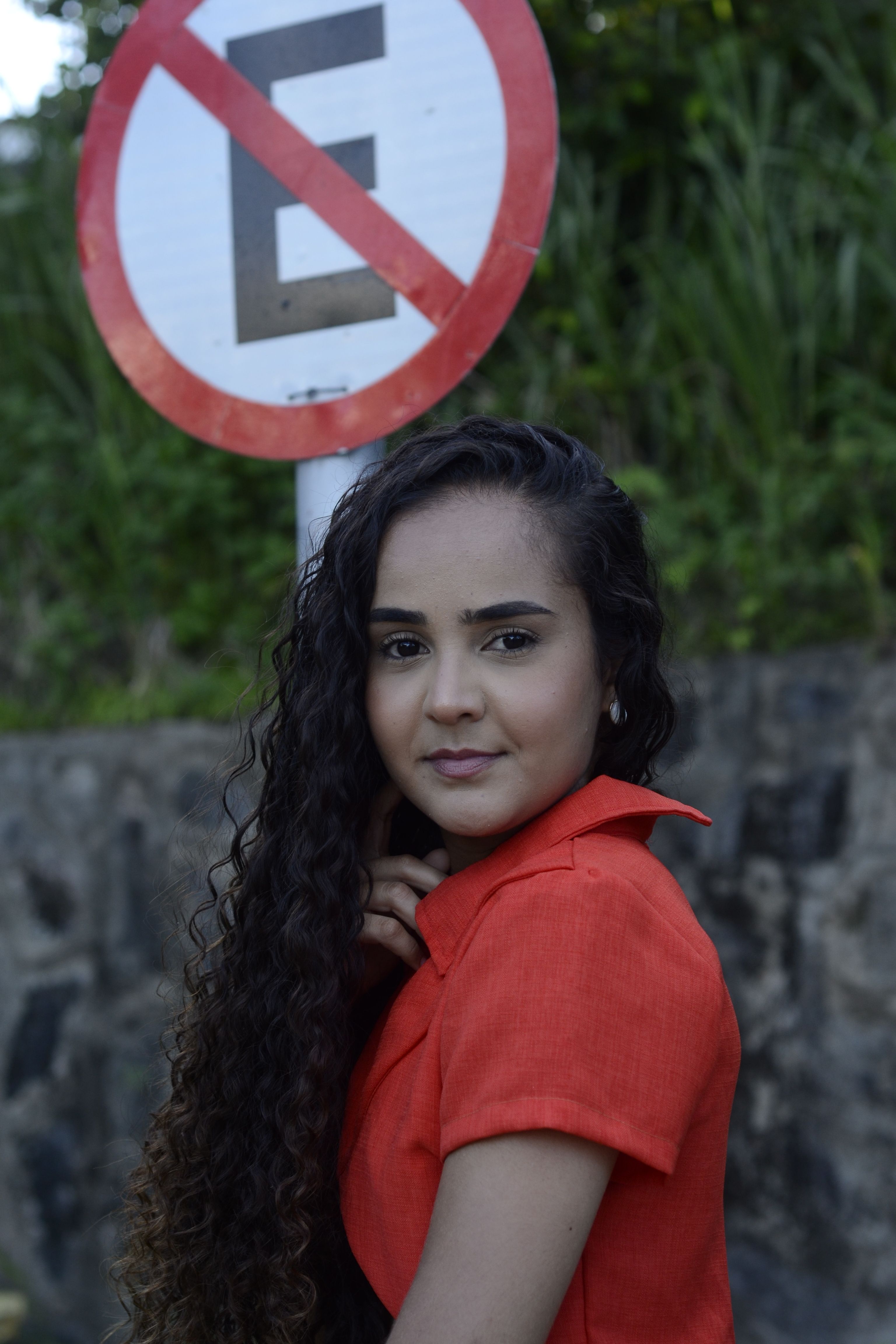 Young woman with curly hair posing near no parking sign outdoors.