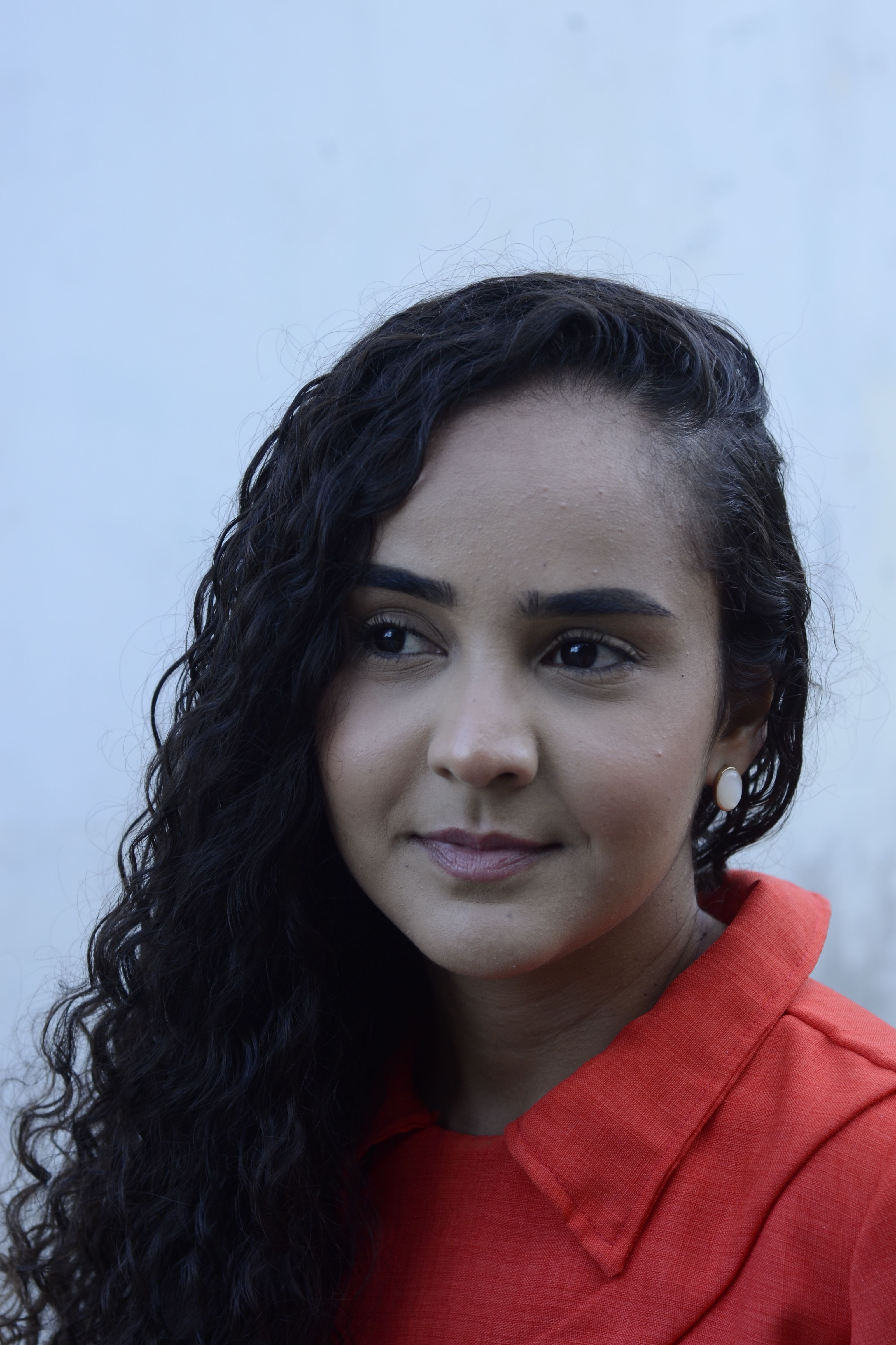 A woman with curly hair smiles softly, wearing a red outfit.