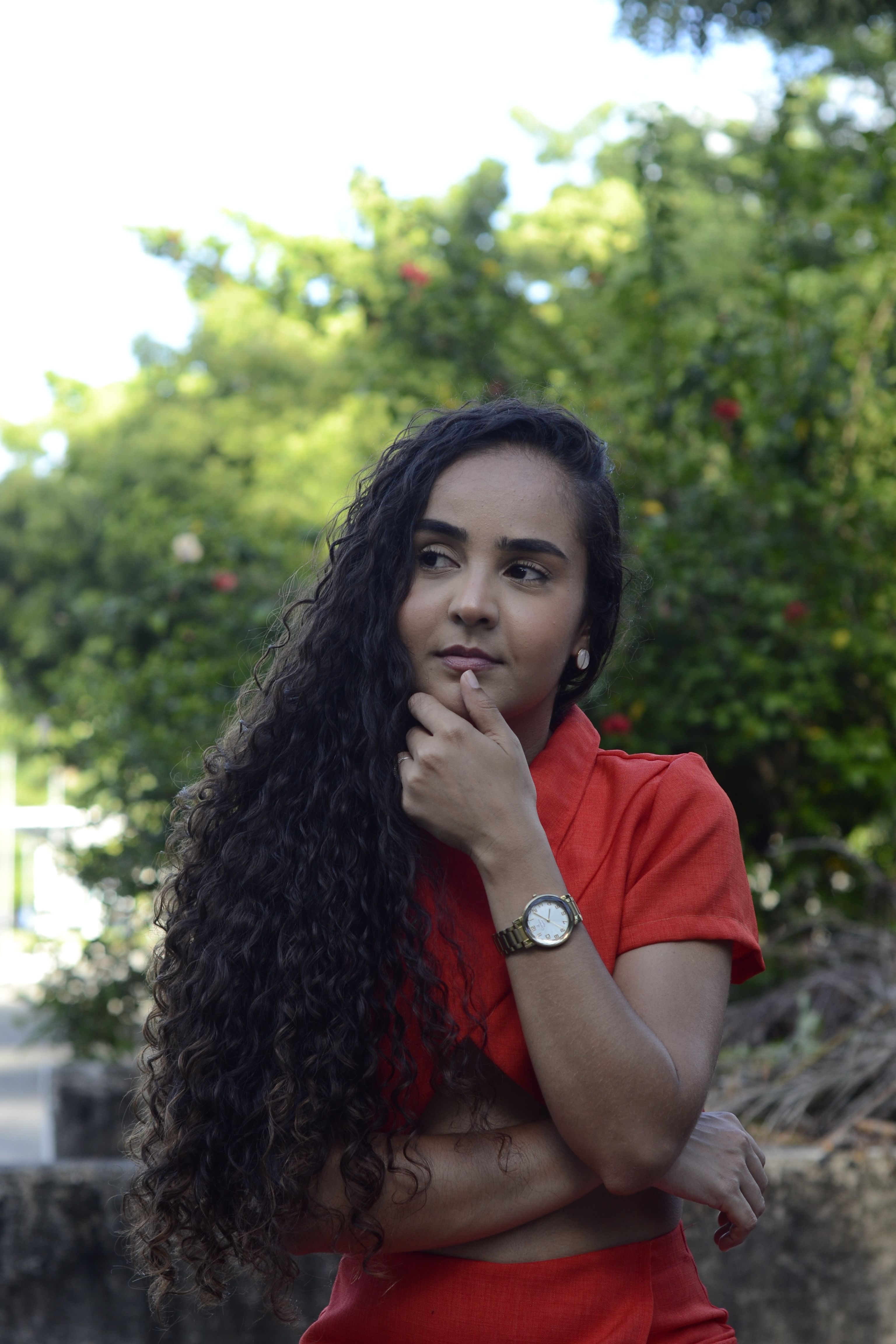 Thoughtful lady in red dress posing outdoors, surrounded by greenery on a bright day.