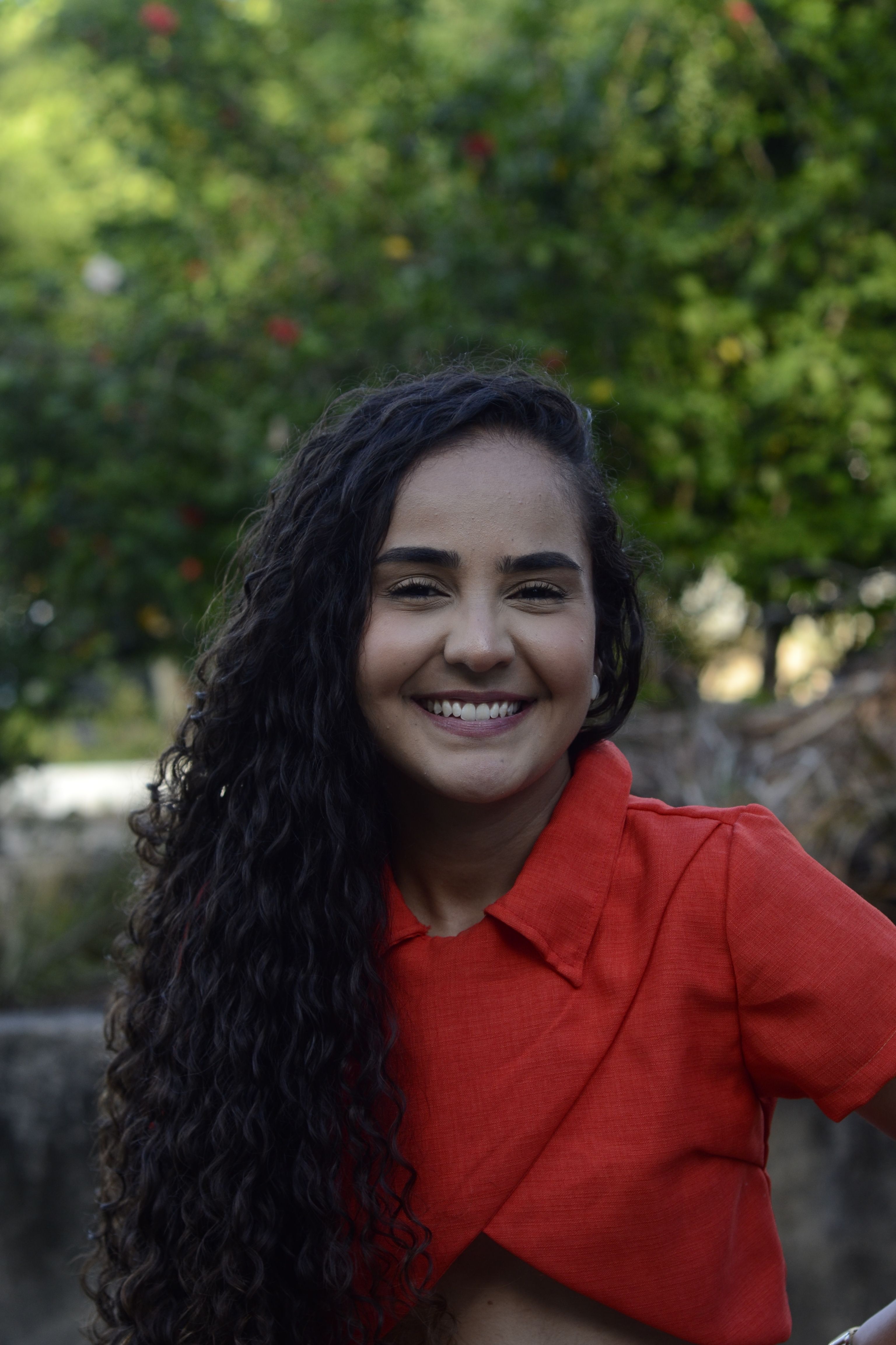 Portrait of a smiling woman with curly hair wearing a red shirt in an outdoor setting.