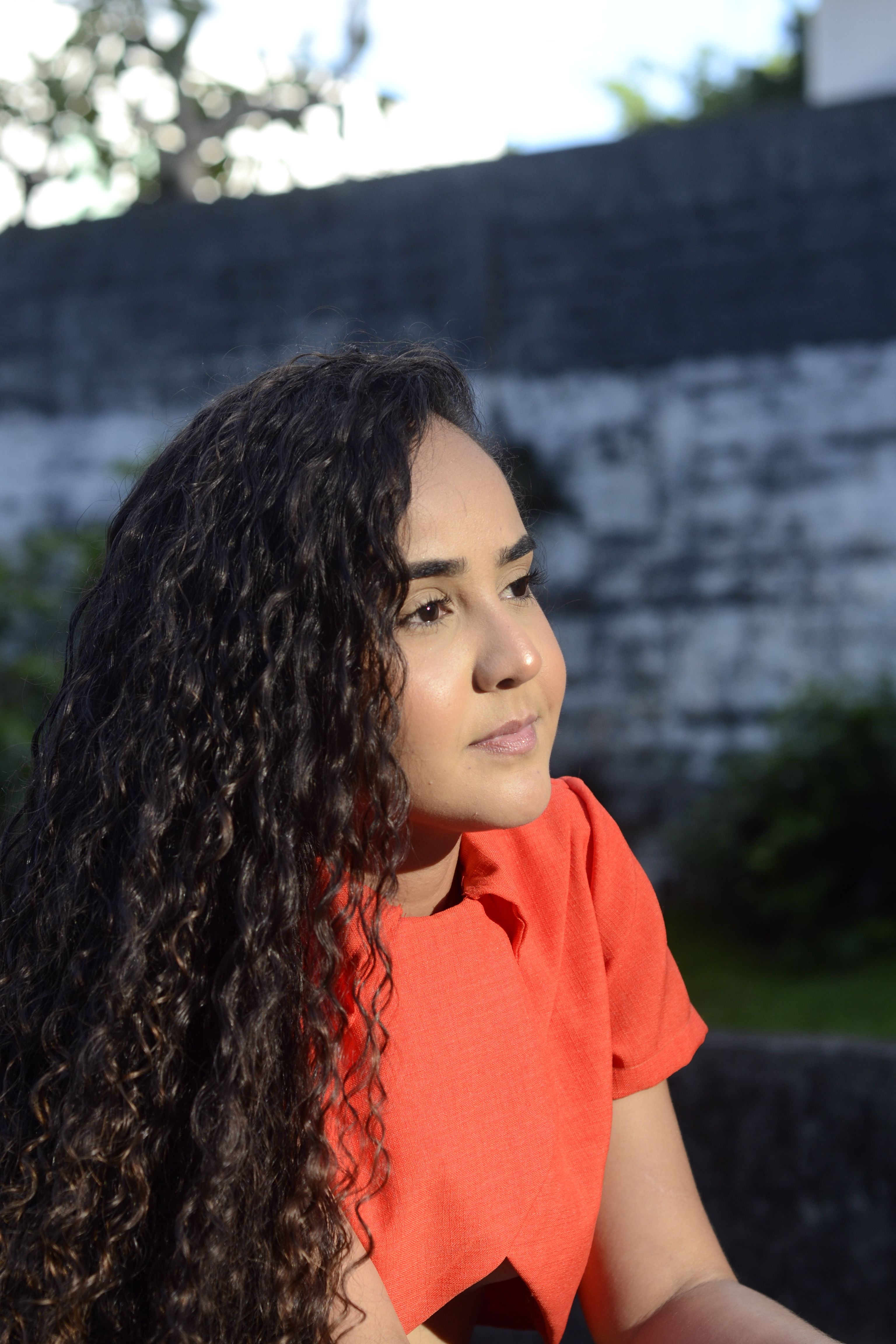 A woman with curly hair wearing a red dress gazing thoughtfully in a serene outdoor setting.