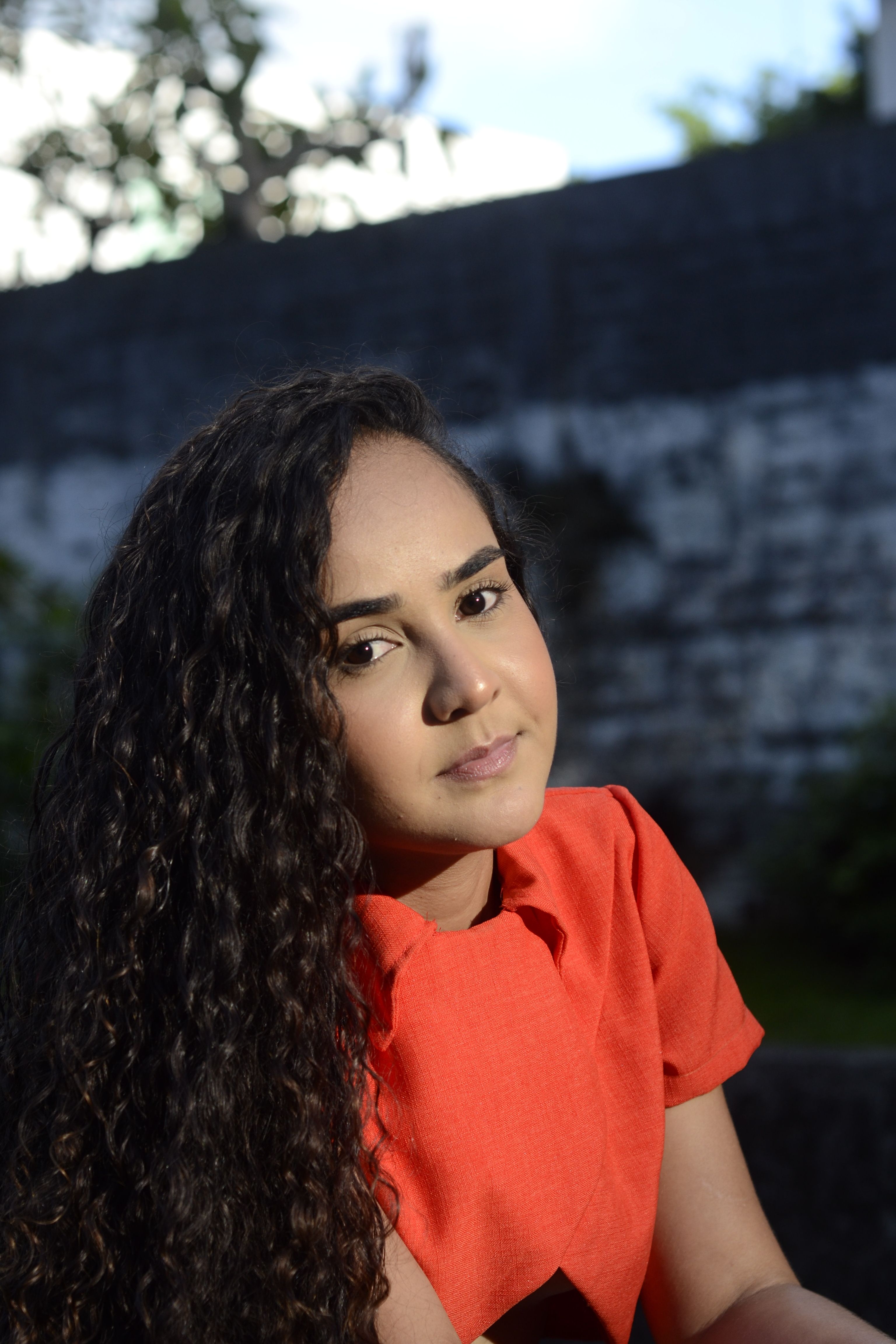A young woman with curly hair, wearing a red dress, seated outdoors.