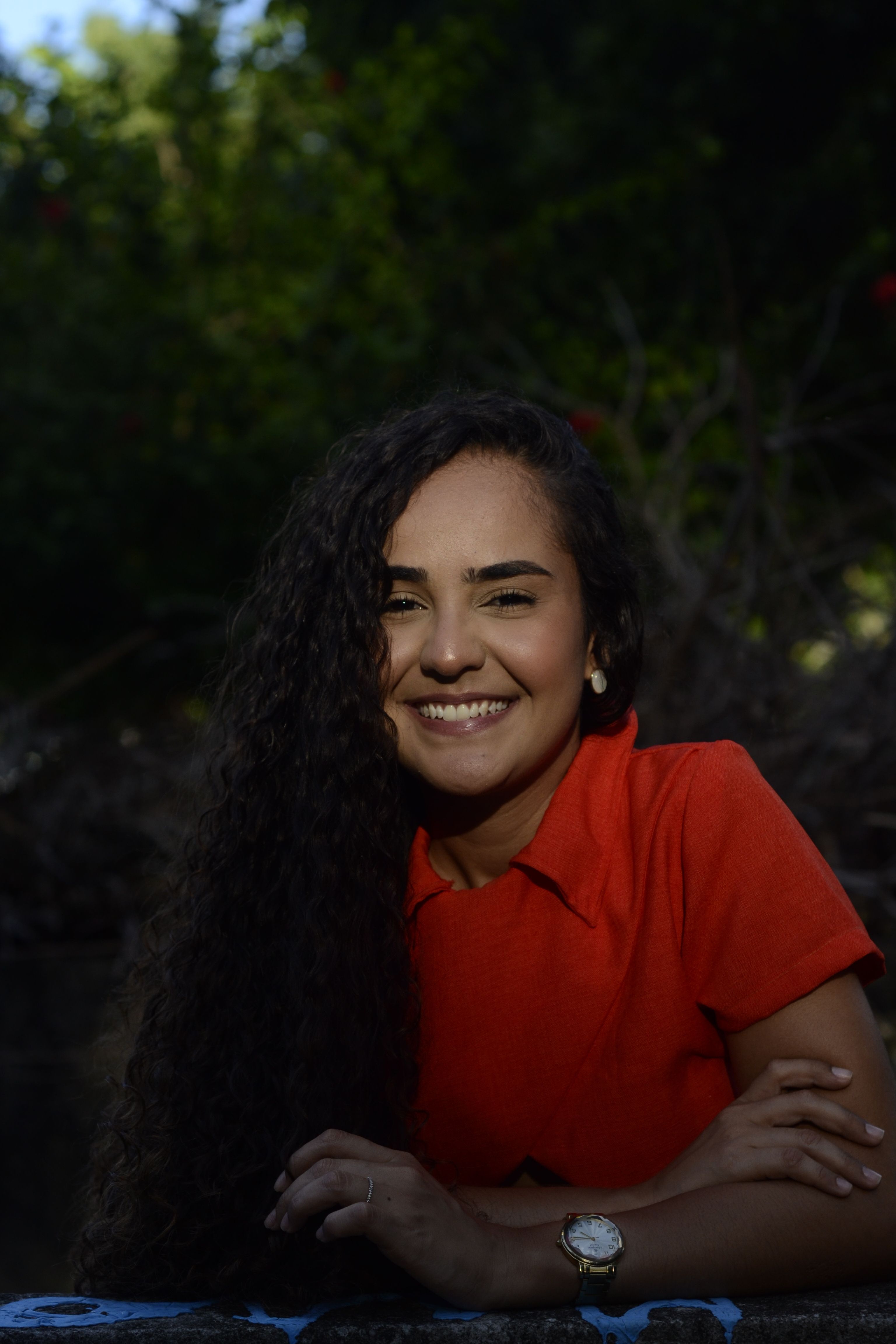 Close-up portrait of a smiling woman in a red outfit outdoors with trees behind.