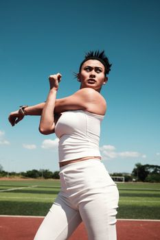A young woman in athletic attire stretching on an outdoor track field under a clear blue sky.