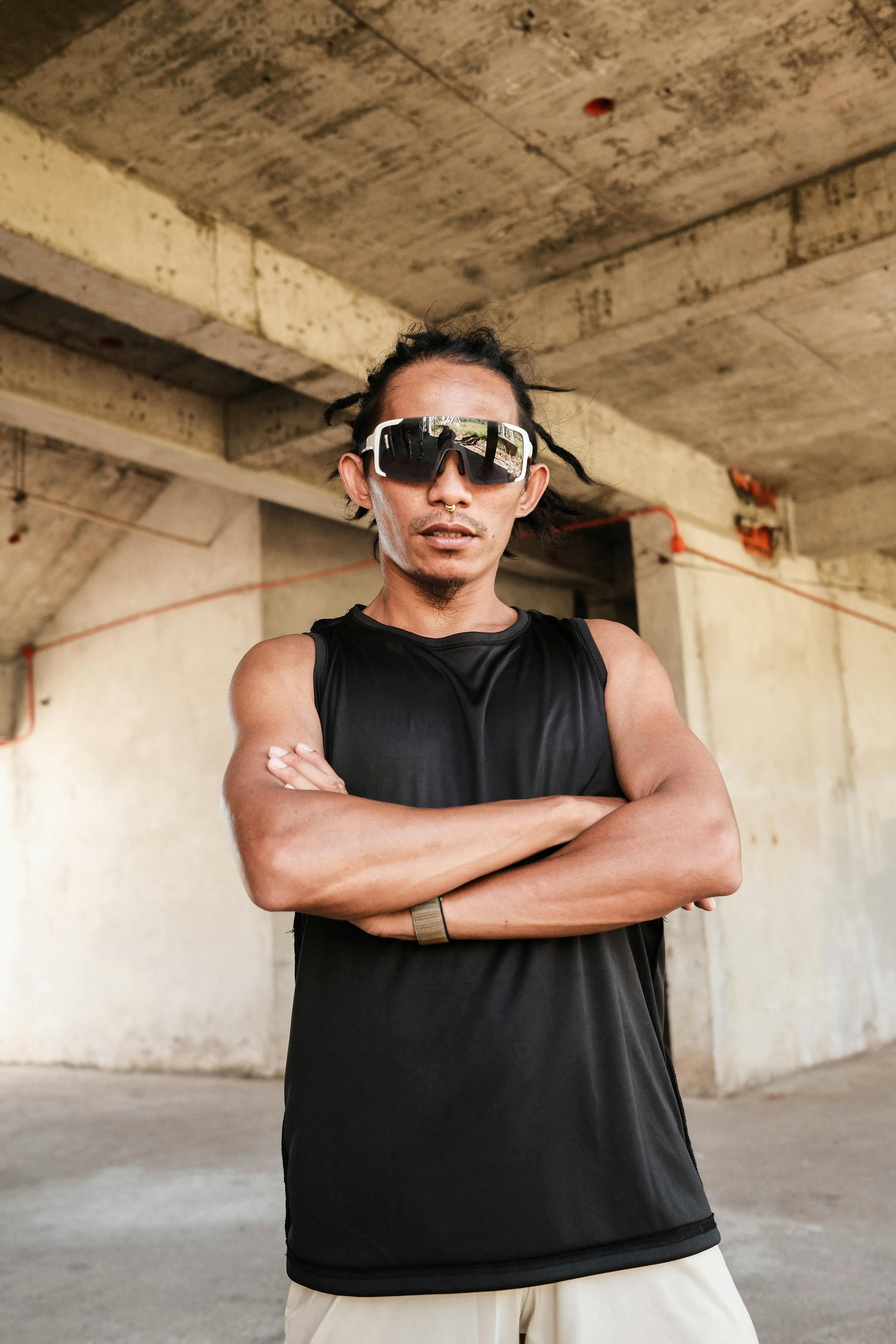 無料 自信に満ちたフィリピン人男性が、サングラスと黒のノースリーブシャツを着て、腕を組んで室内でポーズをとっている。 写真素材