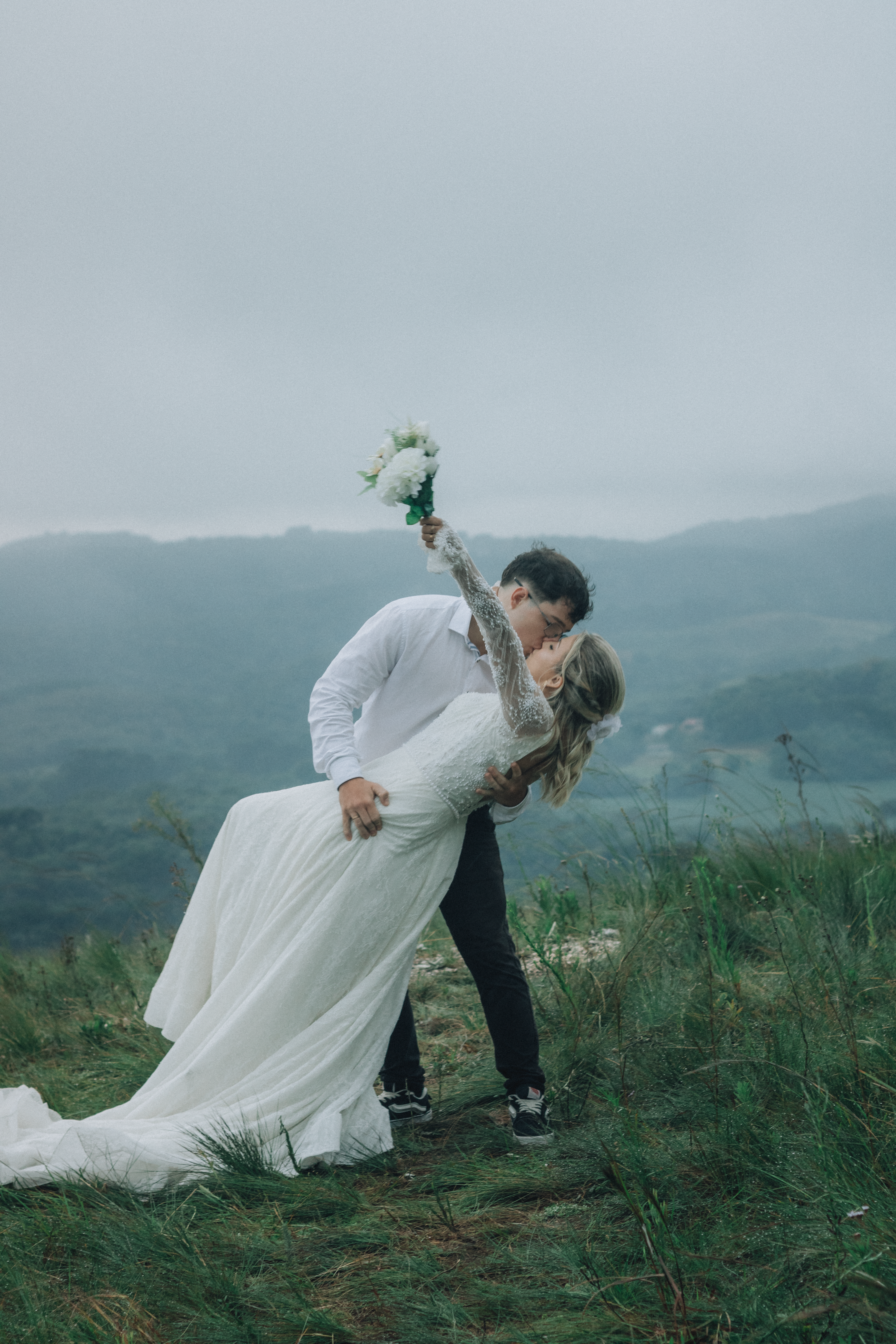 A bride and groom kiss with mountains in the background, holding a bouquet.