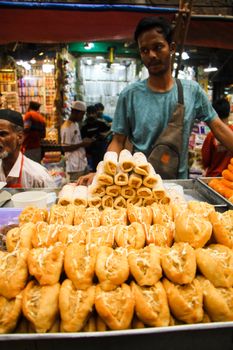 A vibrant street market in Dhaka with a food vendor and fresh pastries on display.