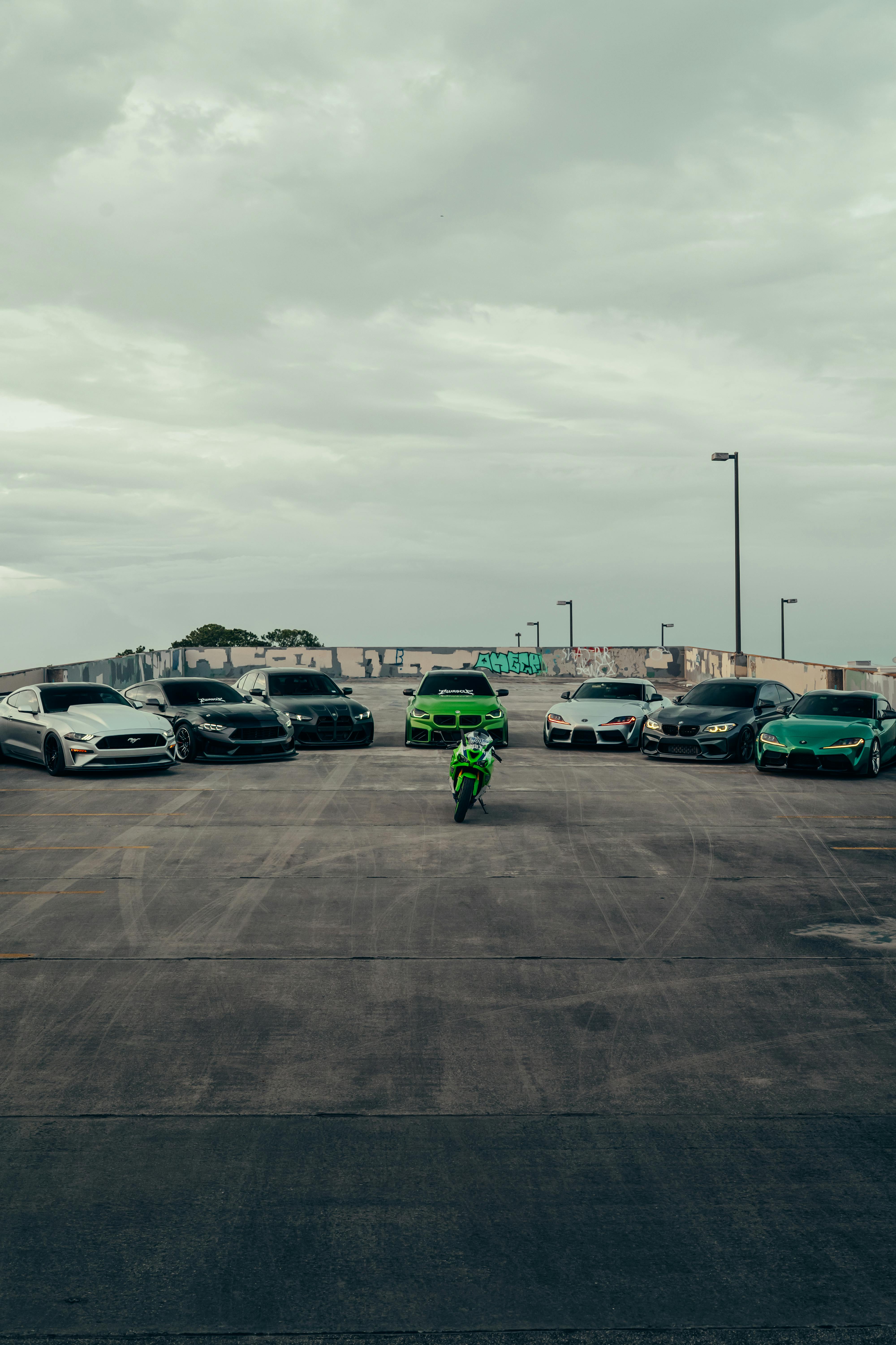 Ücretsiz Bulutlu bir gökyüzü altında, boş bir çatı otoparkında spor arabaların ve yalnız bir motosikletin çarpıcı görüntüsü. Stok Fotoğraflar