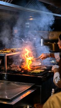 A chefs hands are grilling food with flames rising in a busy restaurant kitchen setting.