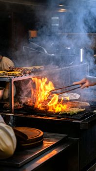 Chef grilling assorted vegetables and meats over open flame in professional kitchen with smoky ambiance.