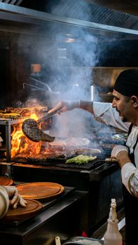 Chef at work, grilling meat and vegetables over an open flame in a busy kitchen.