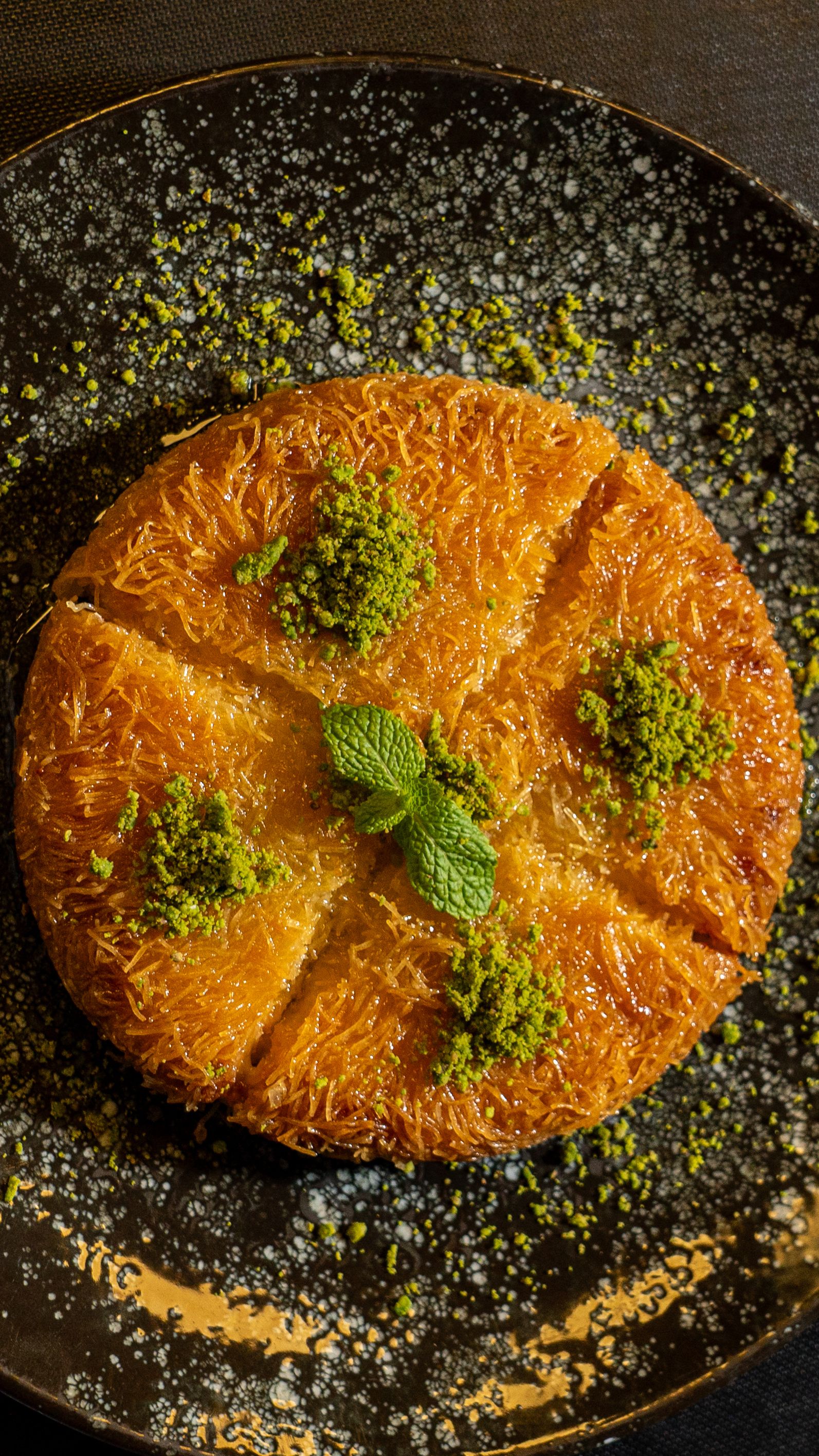 Top view of a sweet kunafa dessert garnished with pistachios and mint leaves, served on a plate.