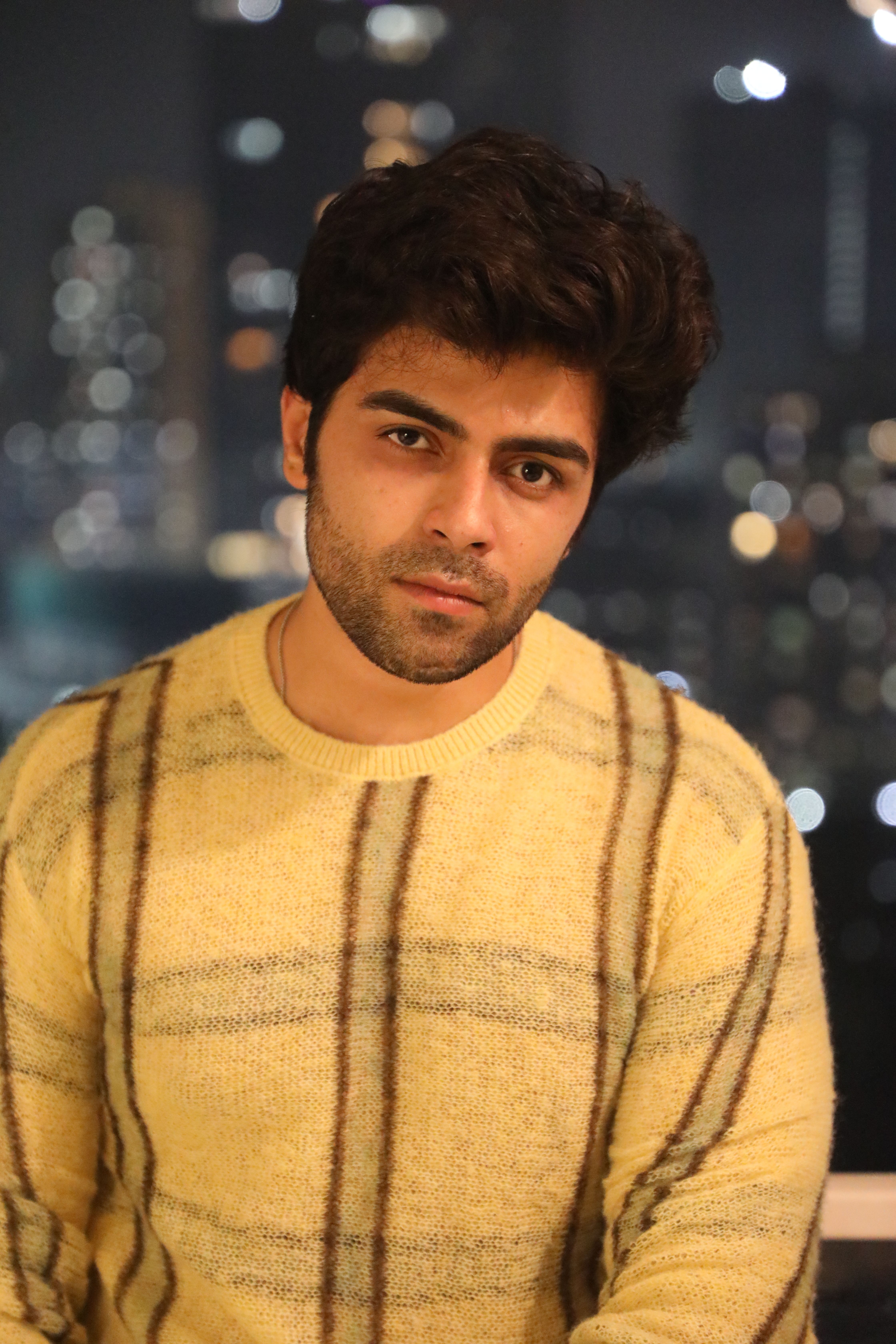 Portrait of a young man against a blurred city night skyline, wearing a cozy sweater.